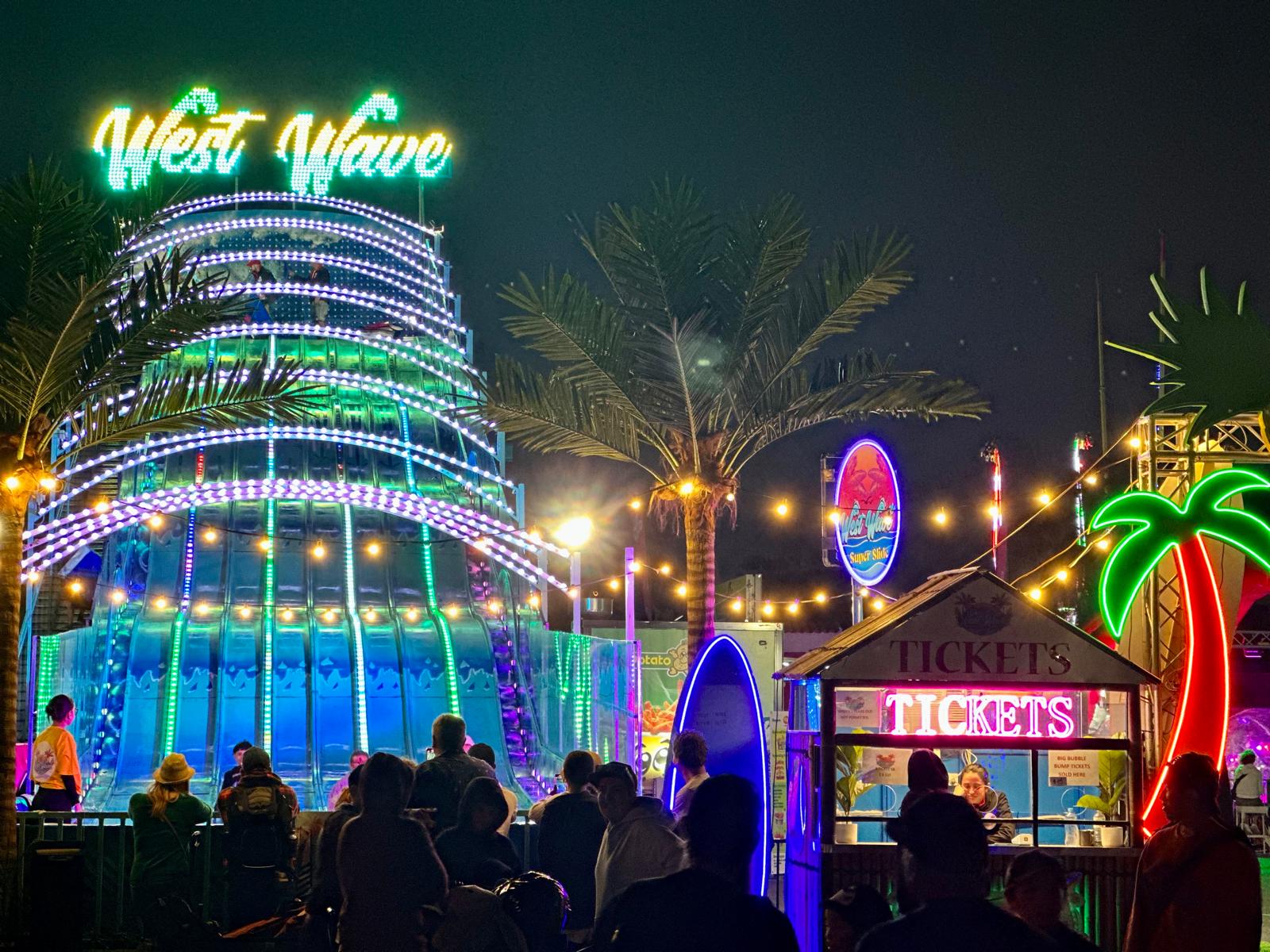 Night scene of an illuminated amusement ride called West Wave with a ticket booth and neon palm tree lights, surrounded by people.