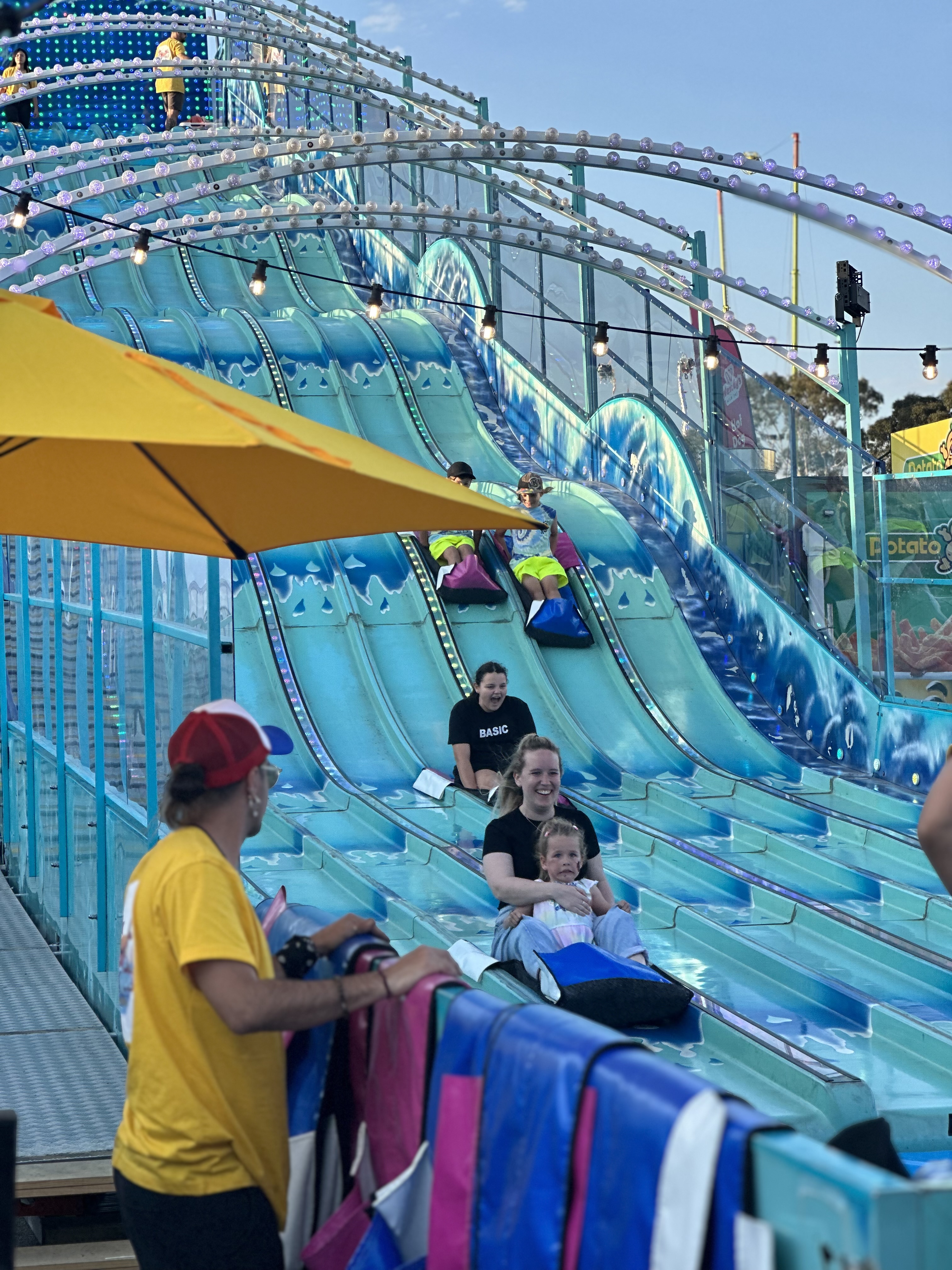 People sliding down a blue multi-lane amusement park slide on mats, with a staff member in a yellow shirt nearby.