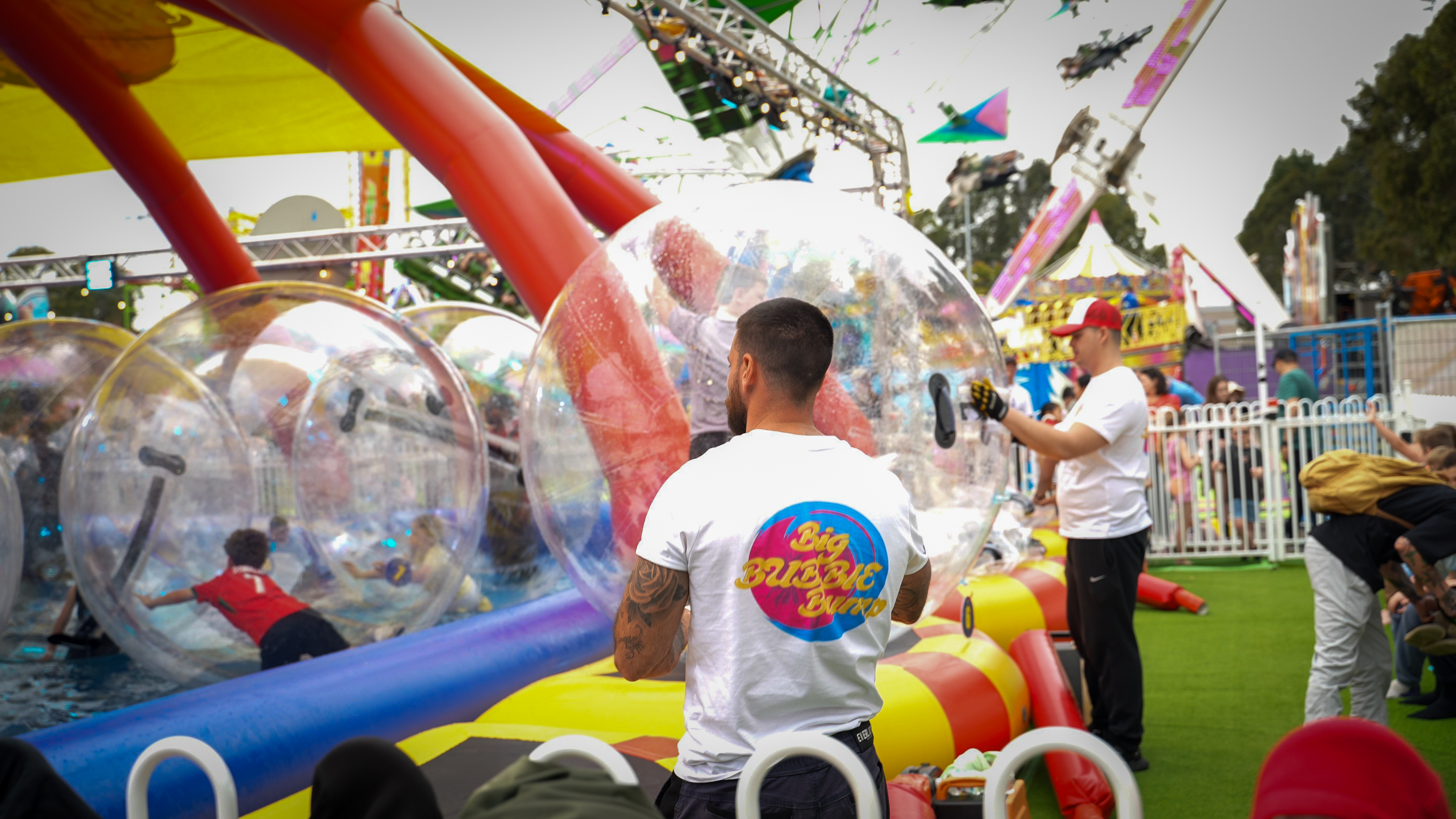 People playing inside large inflatable transparent bubbles at an outdoor amusement park with staff assisting nearby.