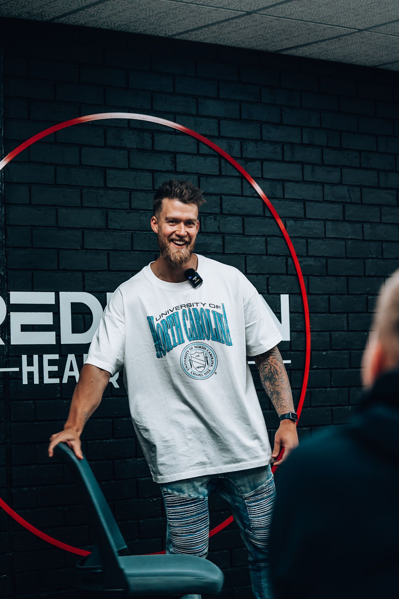 Smiling man with a beard wearing a white University of North Carolina t-shirt, holding a chair in front of a black brick wall with red circle and white text.