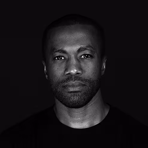 Black and white portrait of a man with a short beard and closely cropped hair wearing a black shirt against a dark background.