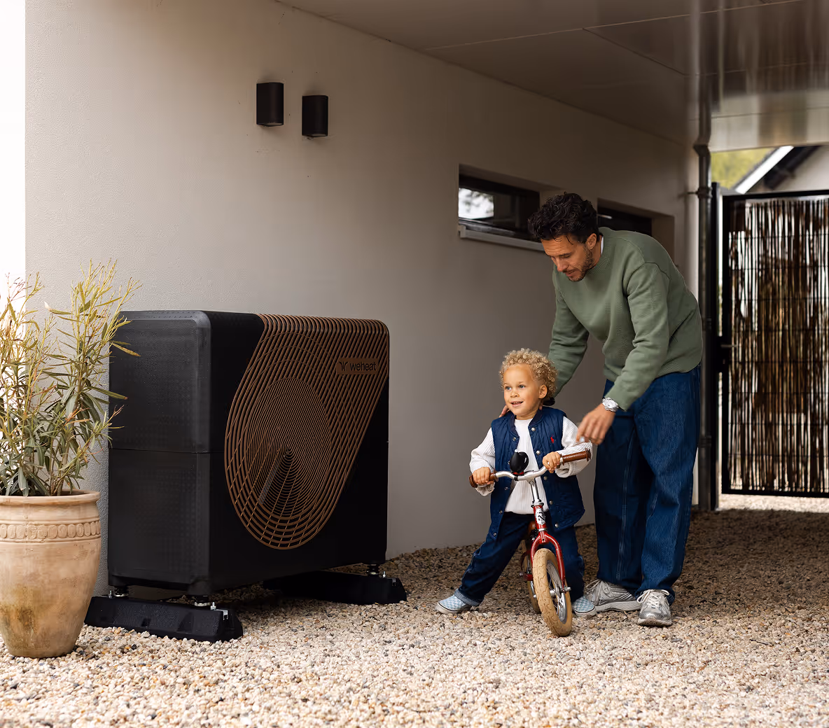 Buitenunit van een warmtepomp naast een woning terwijl een vader zijn kind helpt fietsen.