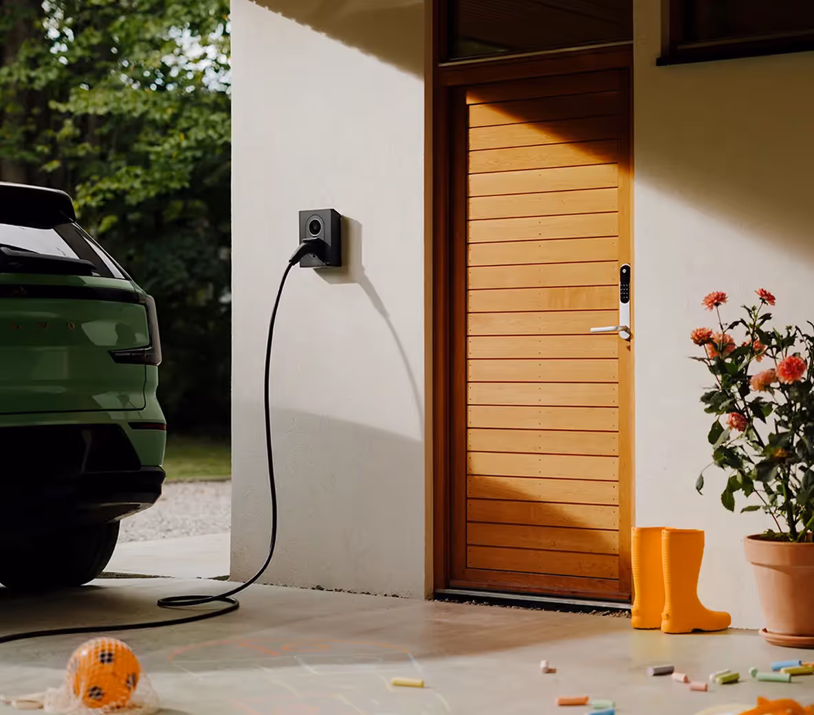 Elektrische auto opgeladen aan een laadpaal bij een moderne woning met houten voordeur en kinderspeelgoed op de oprit.