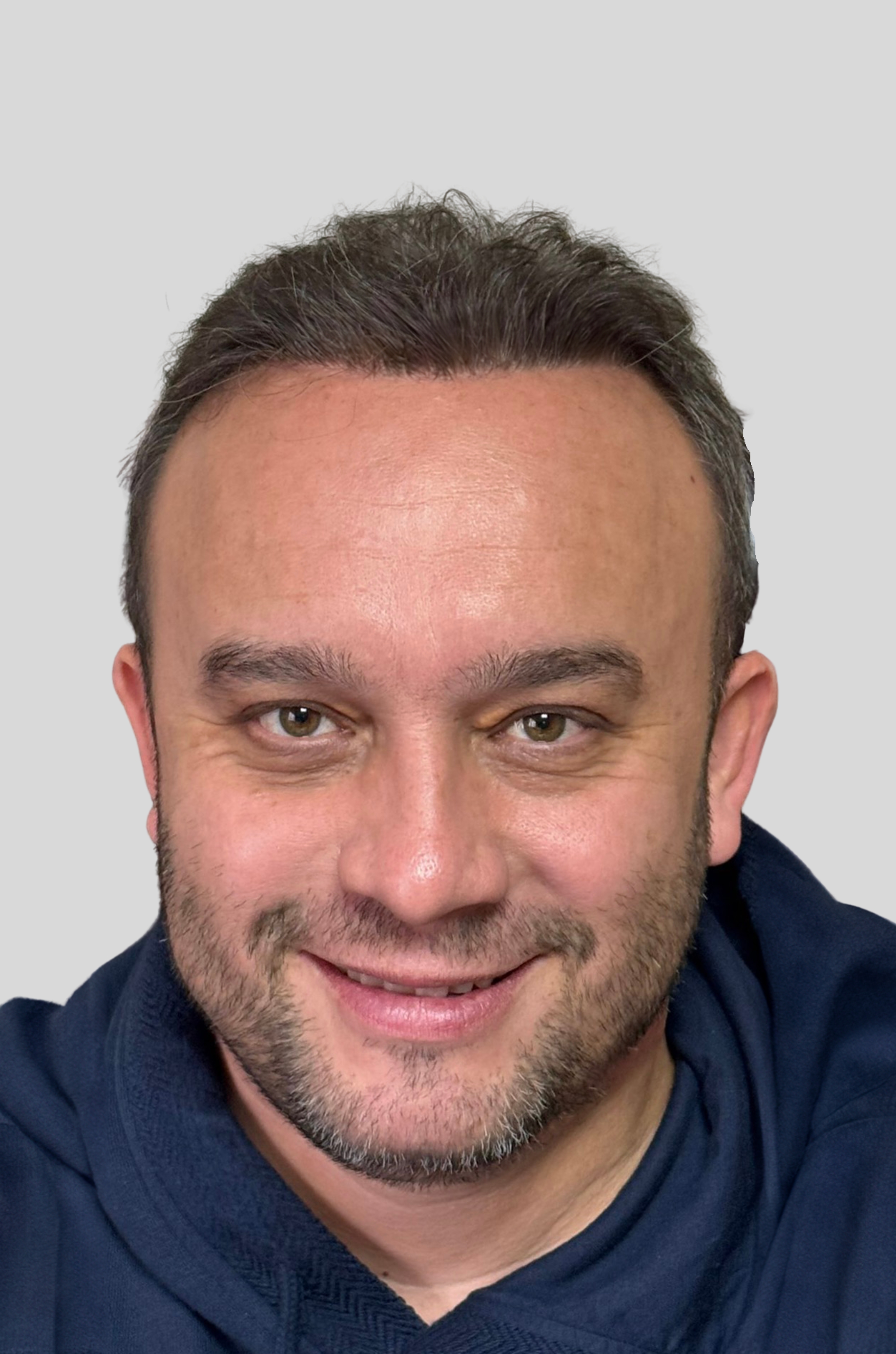 Smiling man with short dark hair, light beard, and wearing a navy blue hoodie against a plain light gray background.