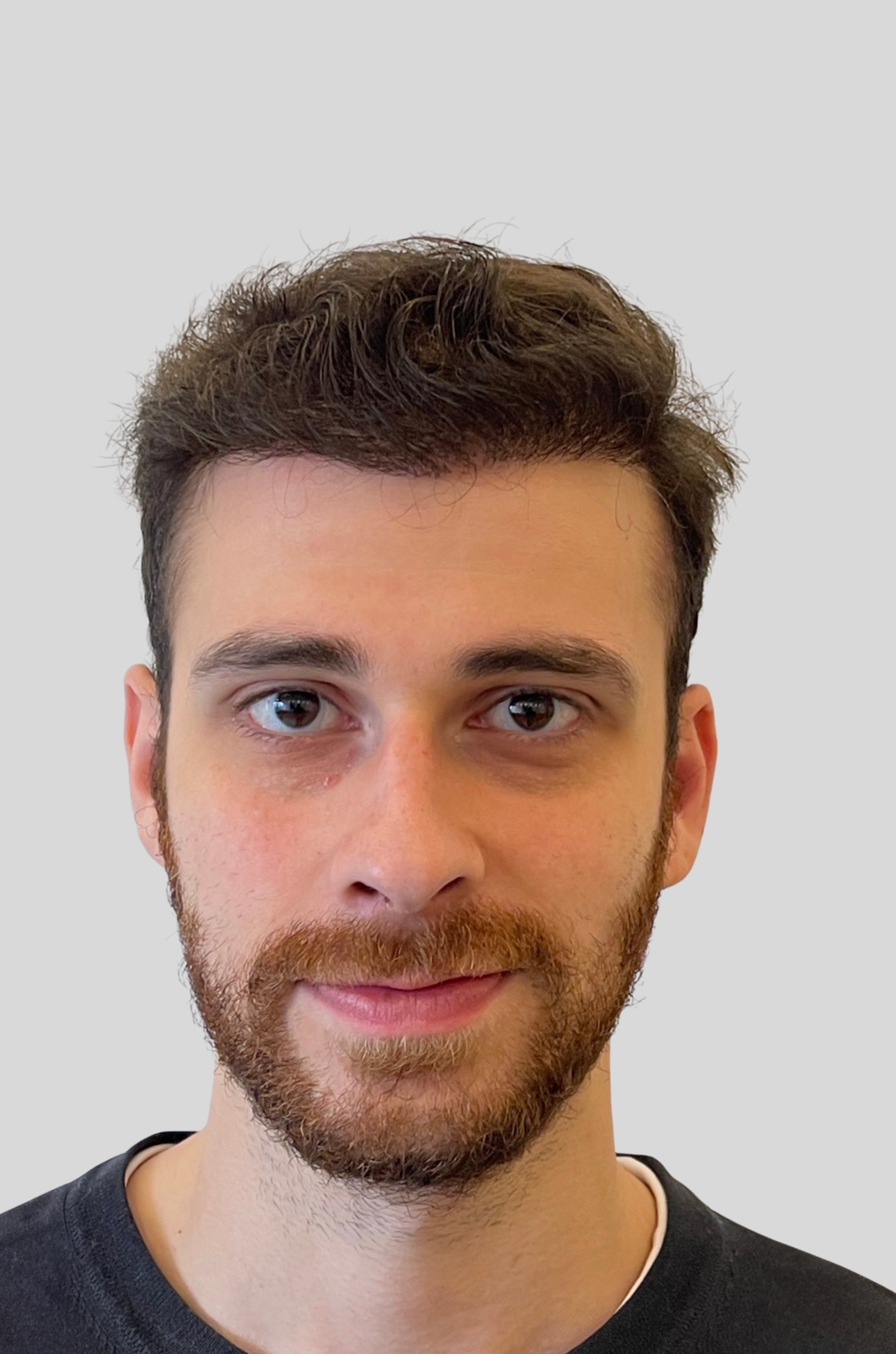 Close-up of a young man with short curly brown hair, a beard, and a slight smile, wearing a dark shirt against a light gray background.