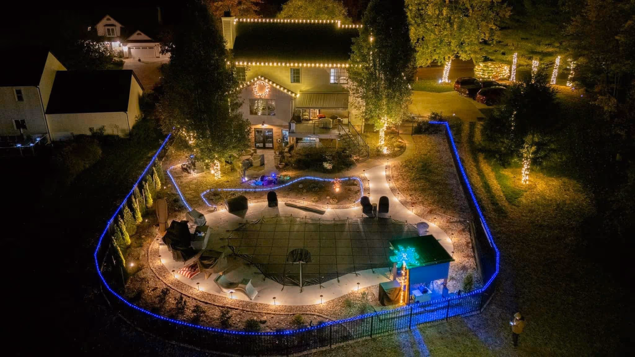 Aerial night view of a backyard with a covered pool, patio furniture, trees wrapped in white string lights, and blue lights outlining the fence.