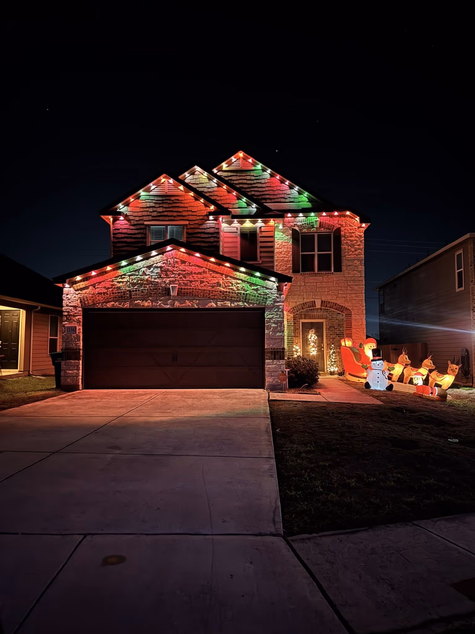 Two-story house decorated with red, green, and white Christmas lights and inflatable Santa, snowman, and reindeer in the front yard at night.