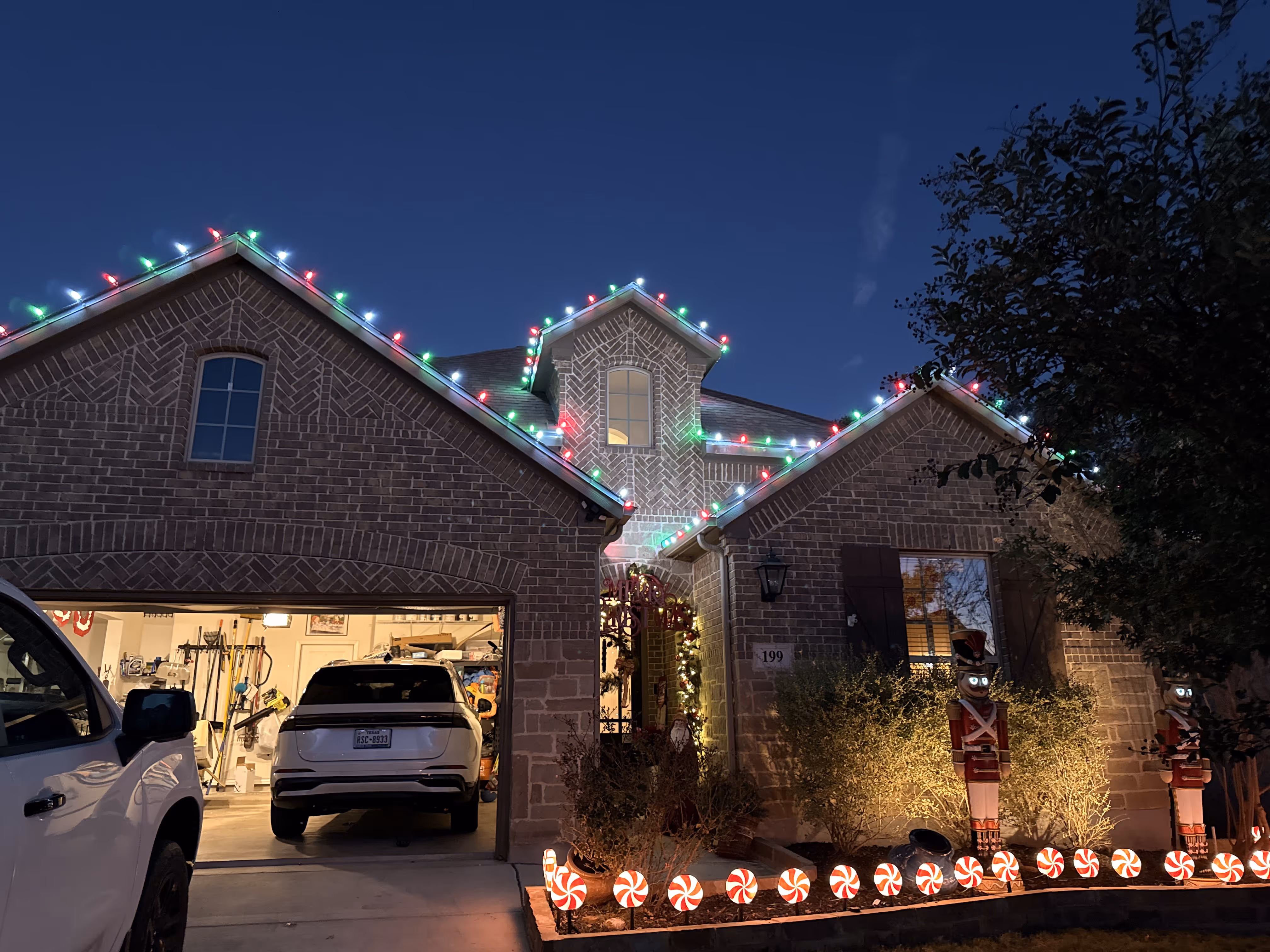 Brick house decorated with multicolored Christmas lights along the roof, gingerbread man lights lining the walkway, and two illuminated nutcracker statues near the front entrance at dusk.