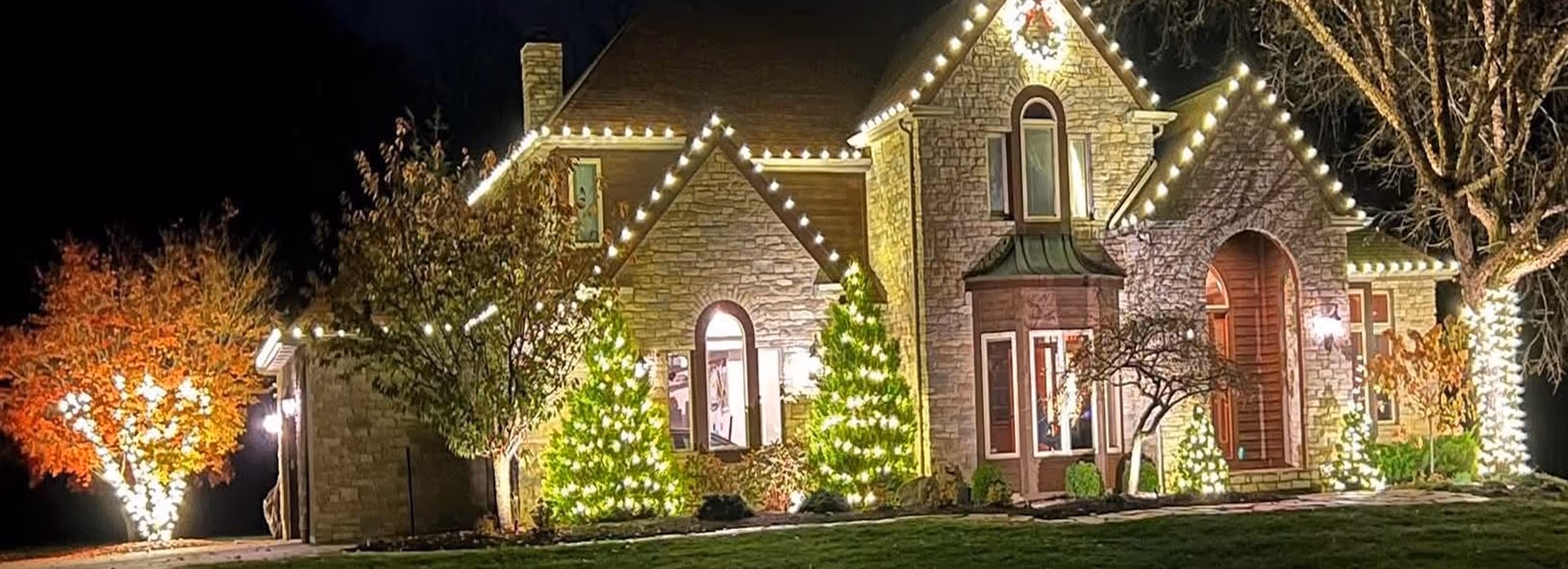 Stone house decorated with white Christmas lights outlining the roof and illuminating trees and bushes in the front yard at night.