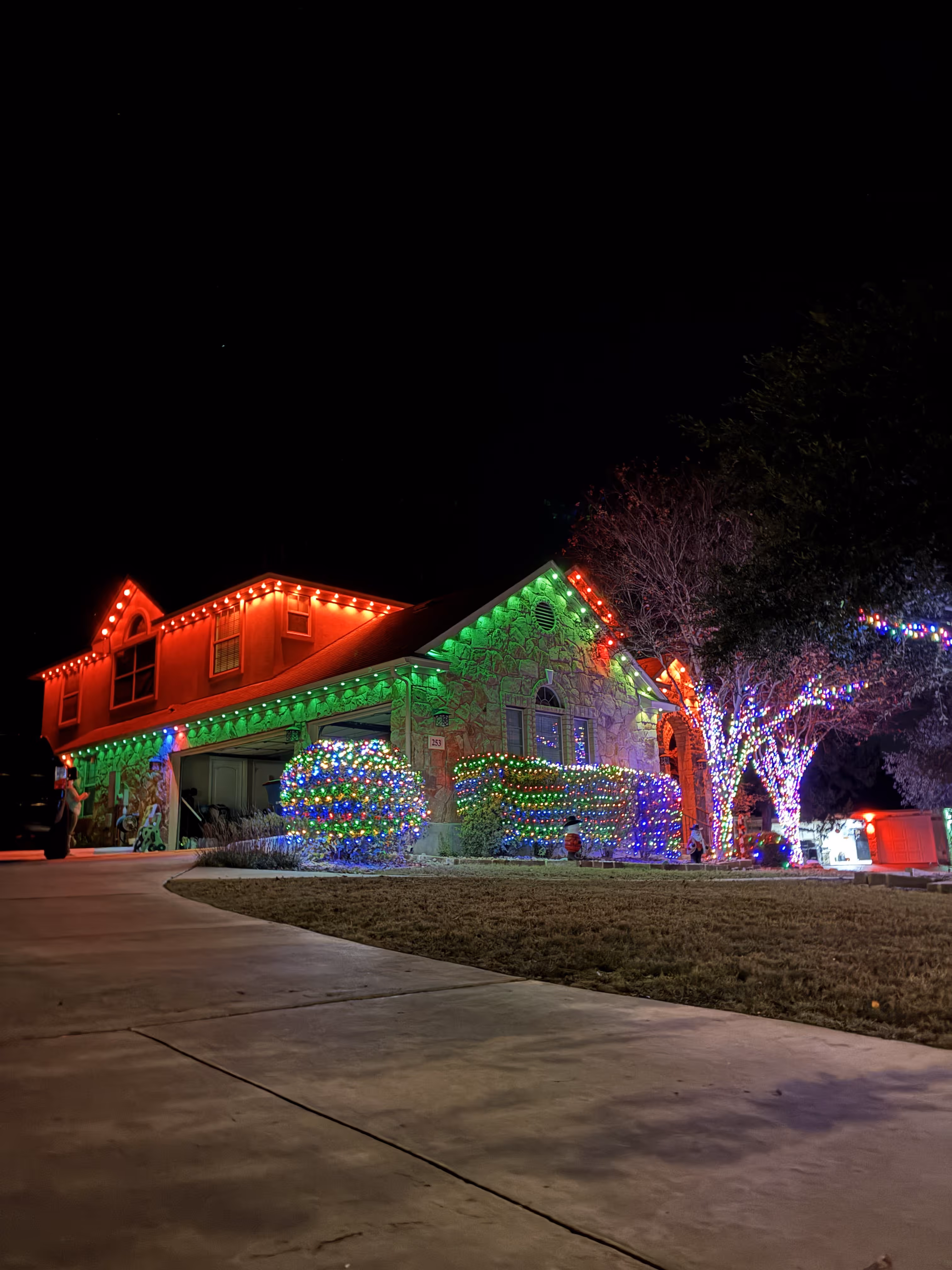 House decorated with colorful Christmas lights on roof edges, bushes, and trees at night.