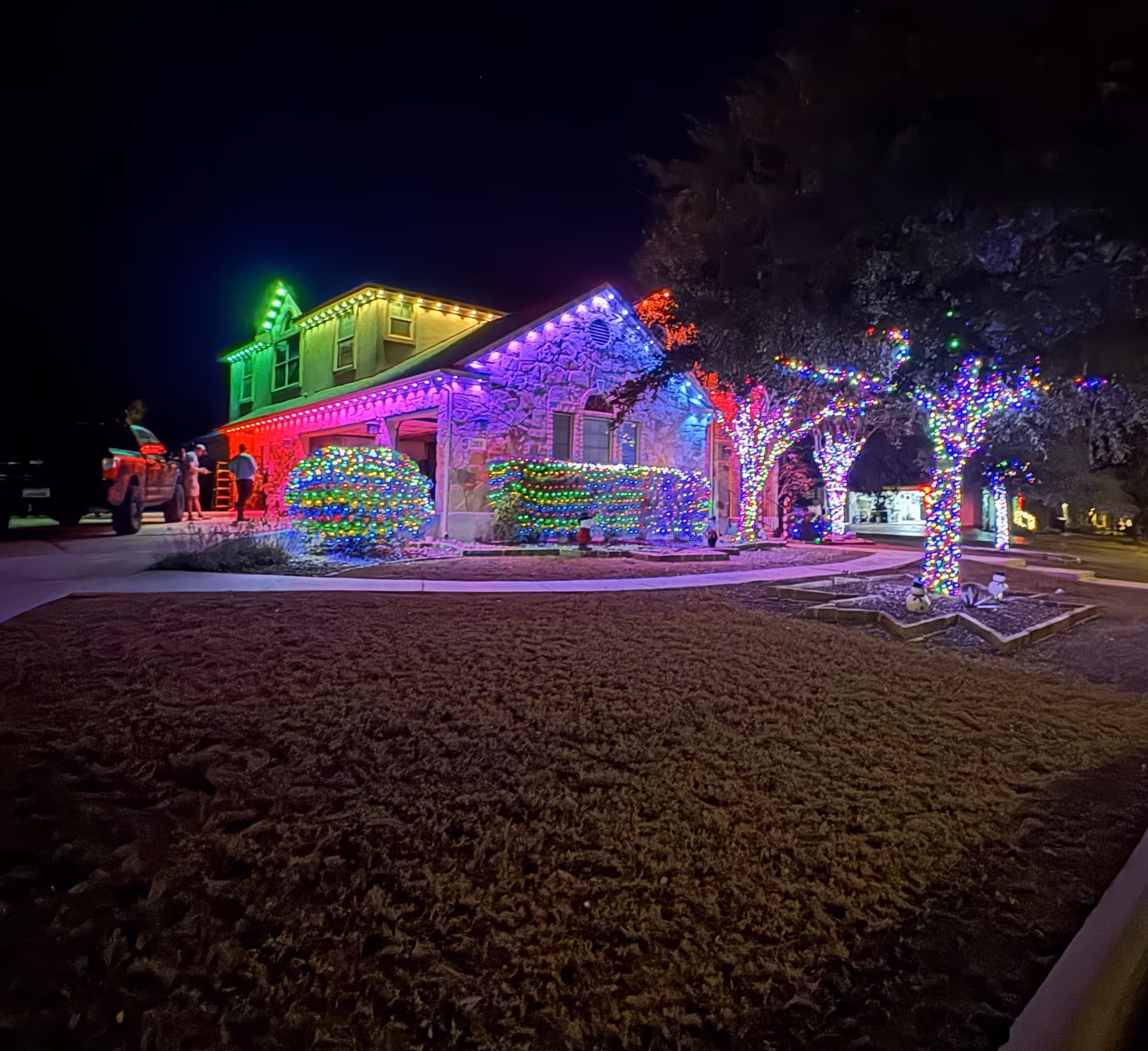 House and trees decorated with multi-colored Christmas lights glowing at night with a truck and people near the driveway.