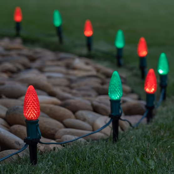 Outdoor pathway lined with alternating red and green Christmas-themed LED lights on stakes in grass beside smooth stones.
