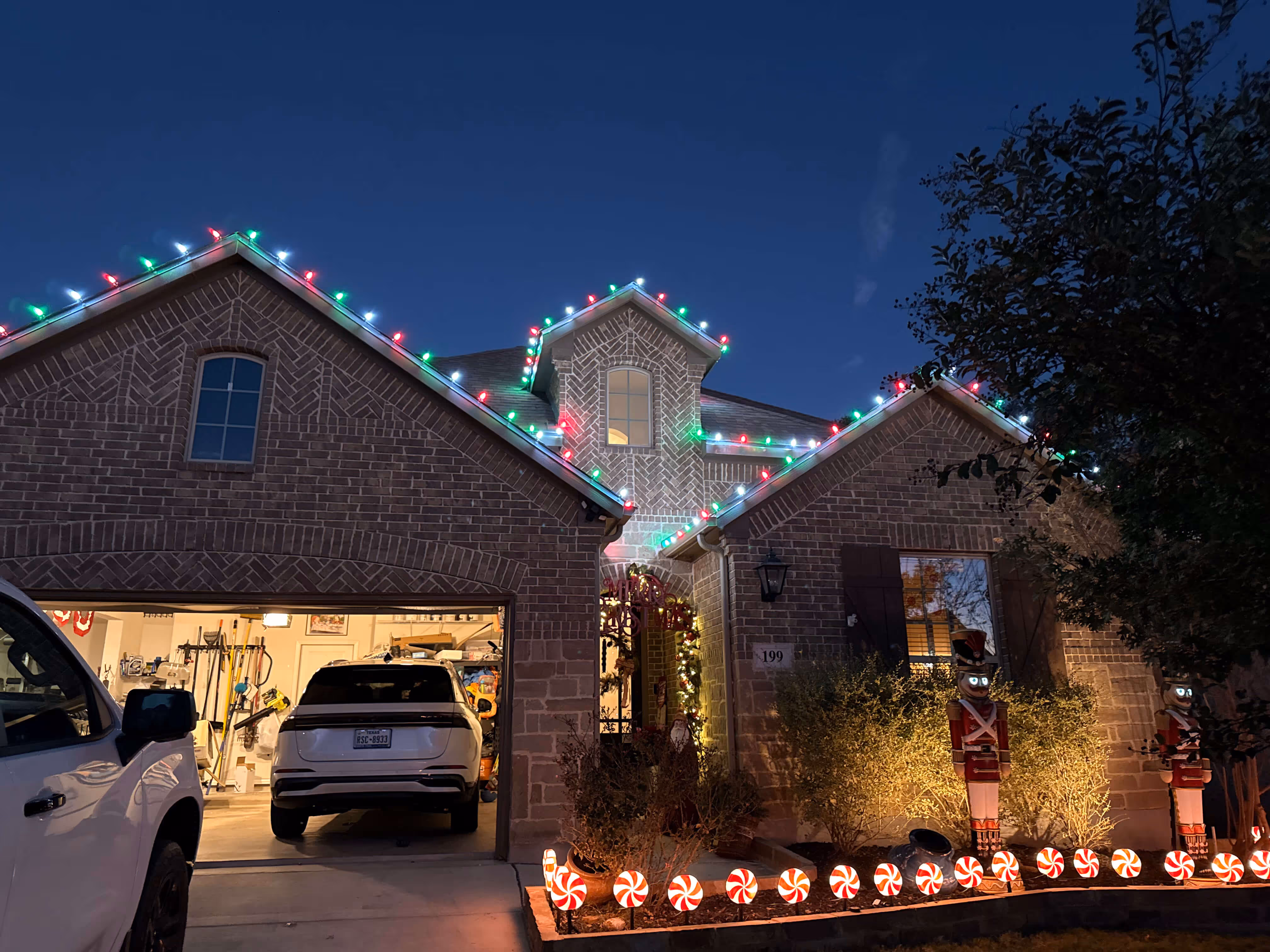 A brick house decorated with colorful Christmas lights along the roof, illuminated nutcracker statues, and candy cane lights lining the front garden at dusk.