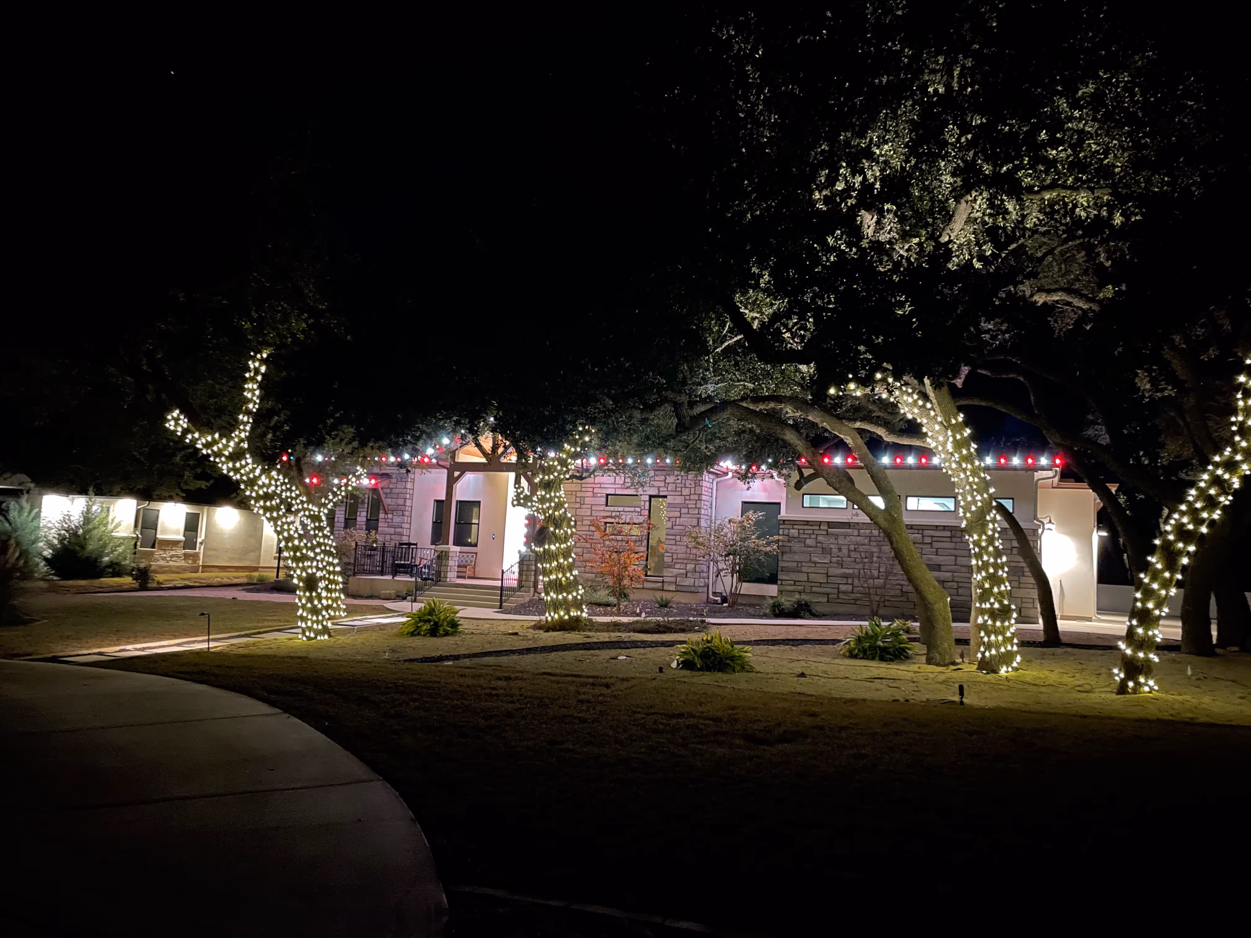 Night view of a house with trees wrapped in white string lights and red and white lights along the roofline.