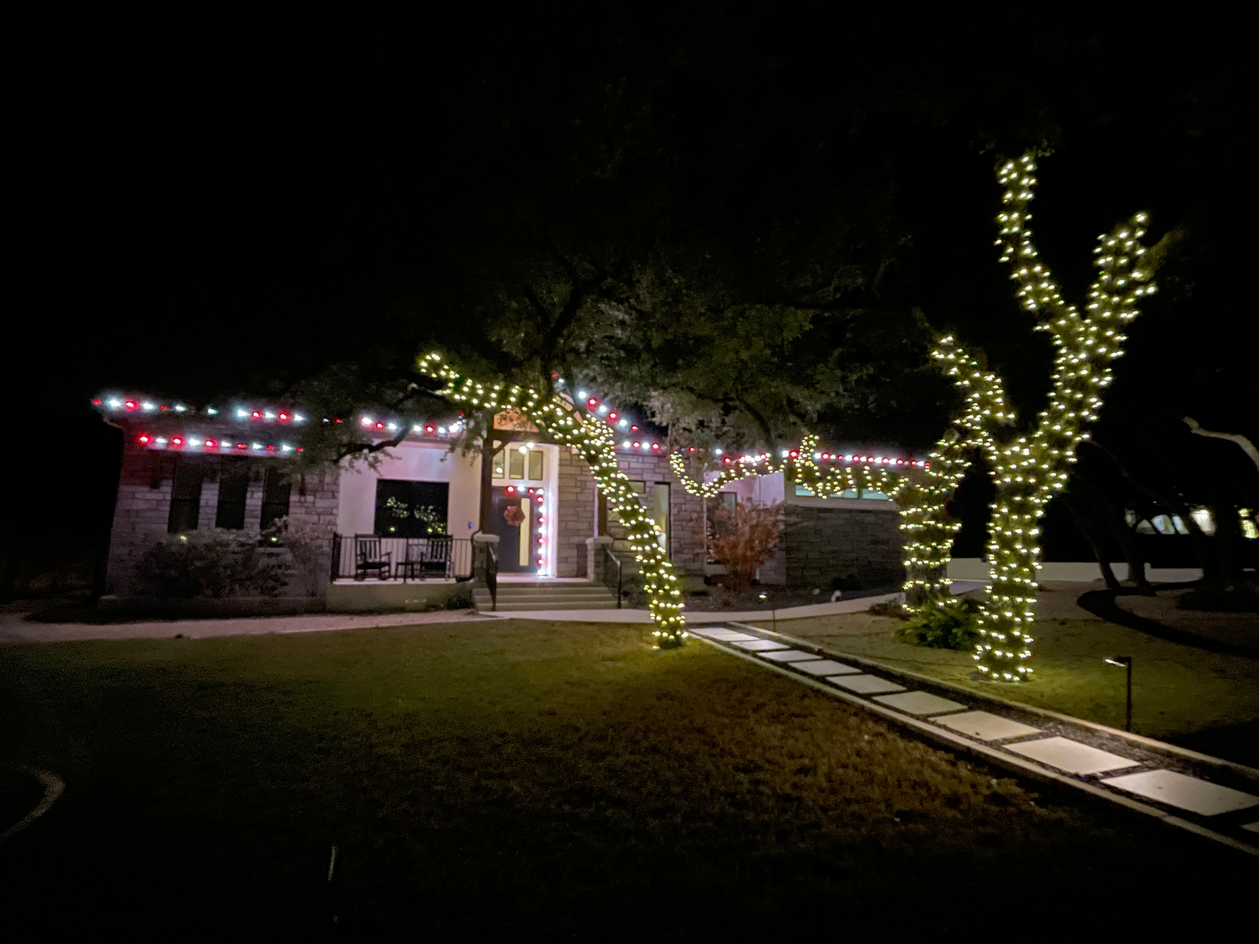 House at night decorated with white and red Christmas lights on the roofline, door, and wrapped around tree trunks.