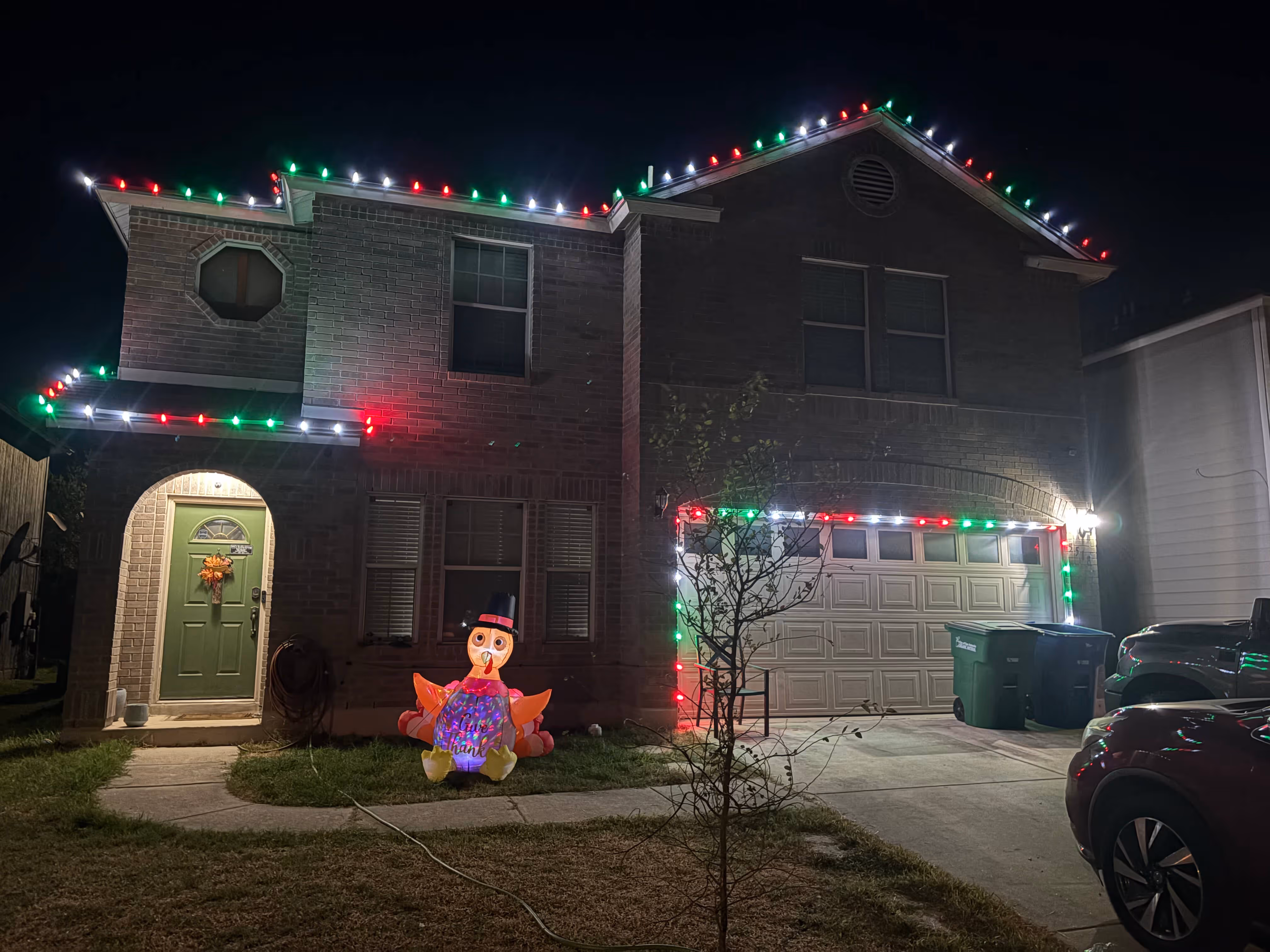 Two-story brick house decorated with red, green, and white Christmas lights along the roofline and garage, with an inflatable Thanksgiving turkey on the front lawn at night.