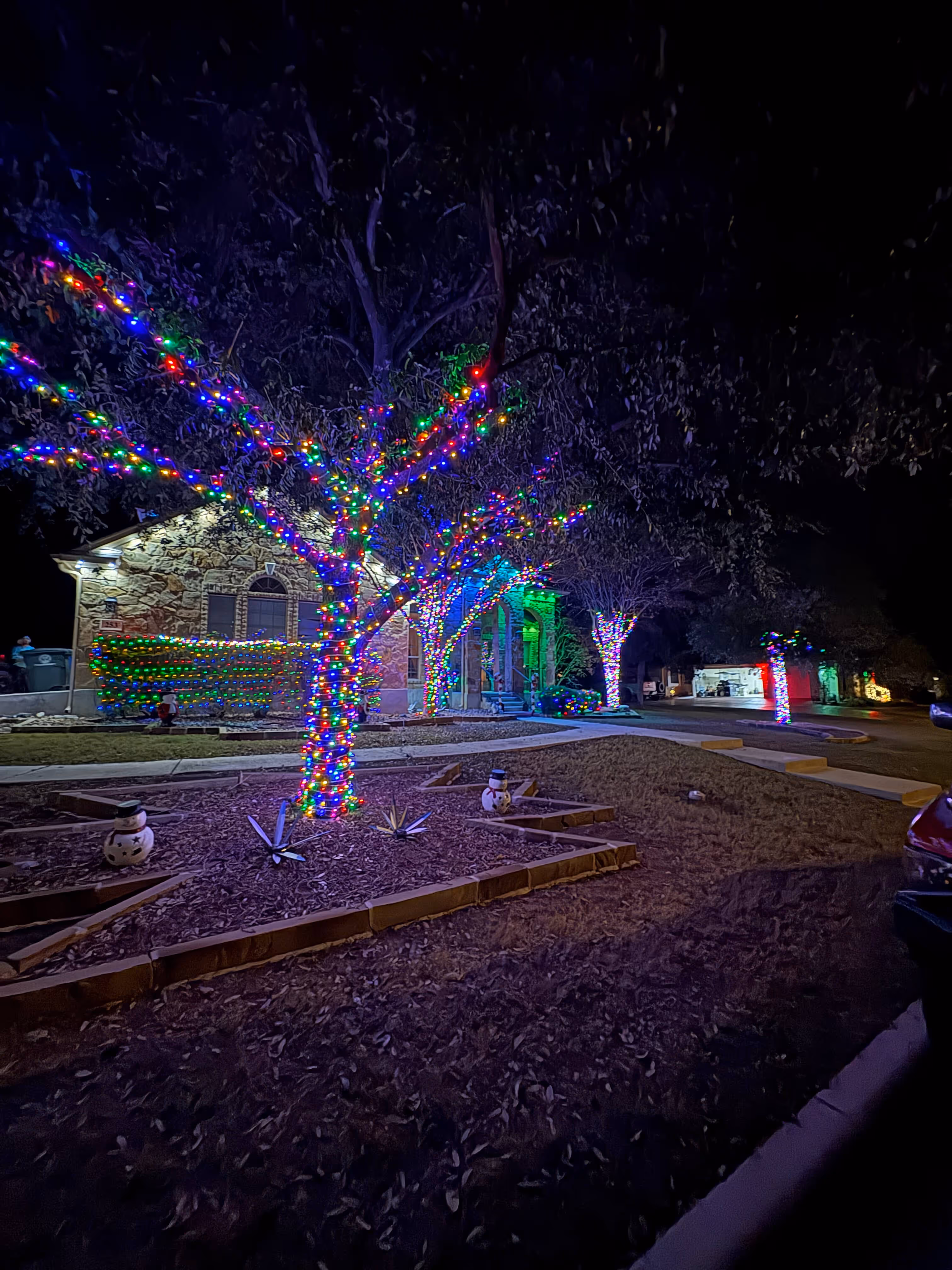 Nighttime view of a house with multiple trees wrapped in colorful Christmas lights and small snowman decorations on the ground.