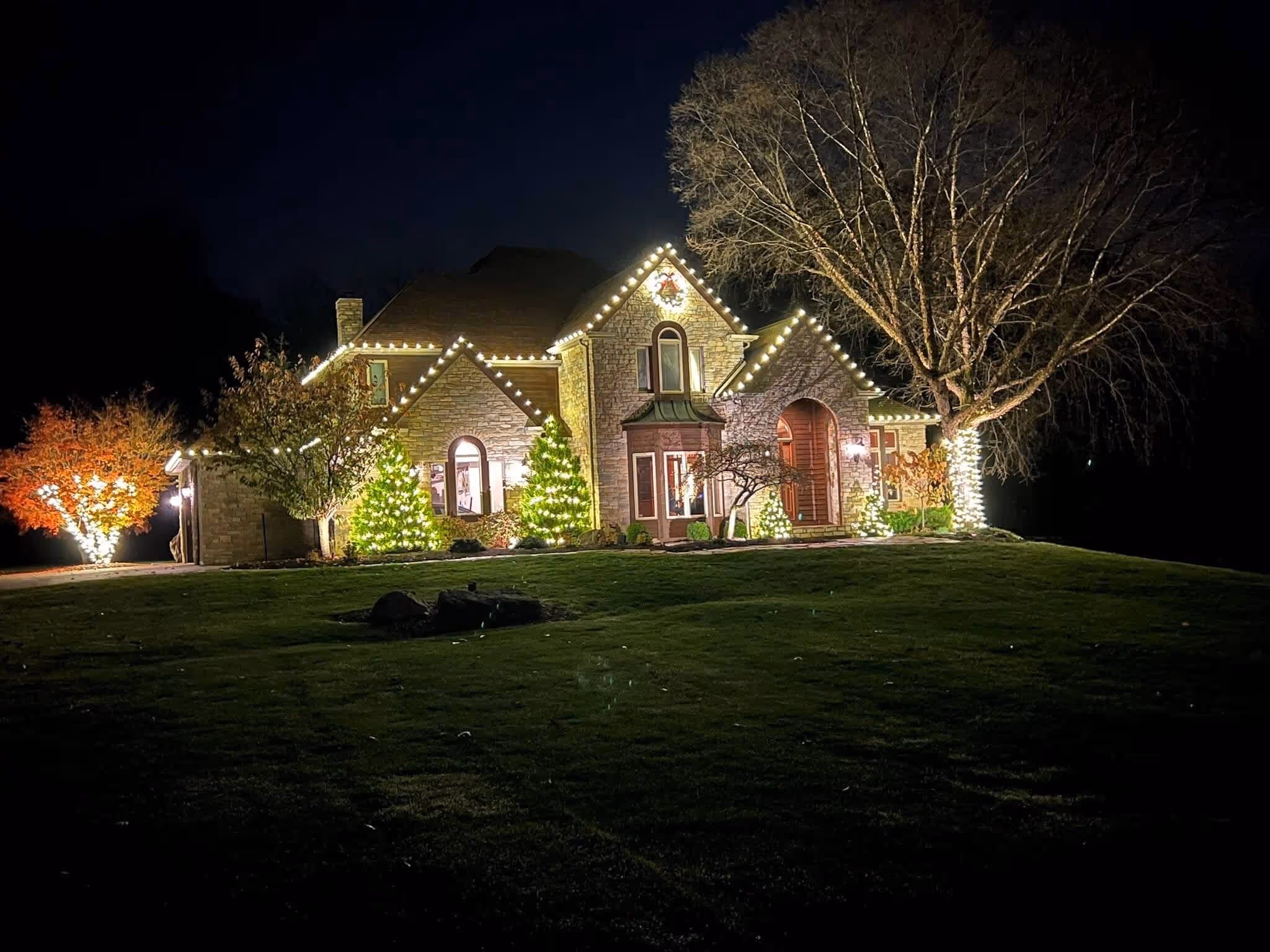 Stone house at night decorated with white Christmas lights outlining roof edges, trees, and bushes.