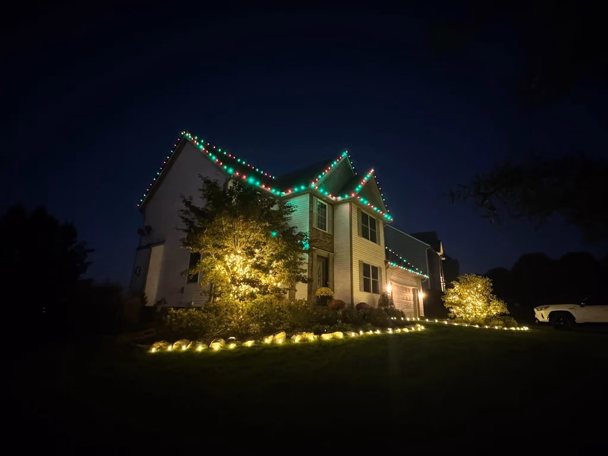 Two-story house at night decorated with red and green Christmas lights along the roof and warm white lights illuminating shrubs and lining the garden border.