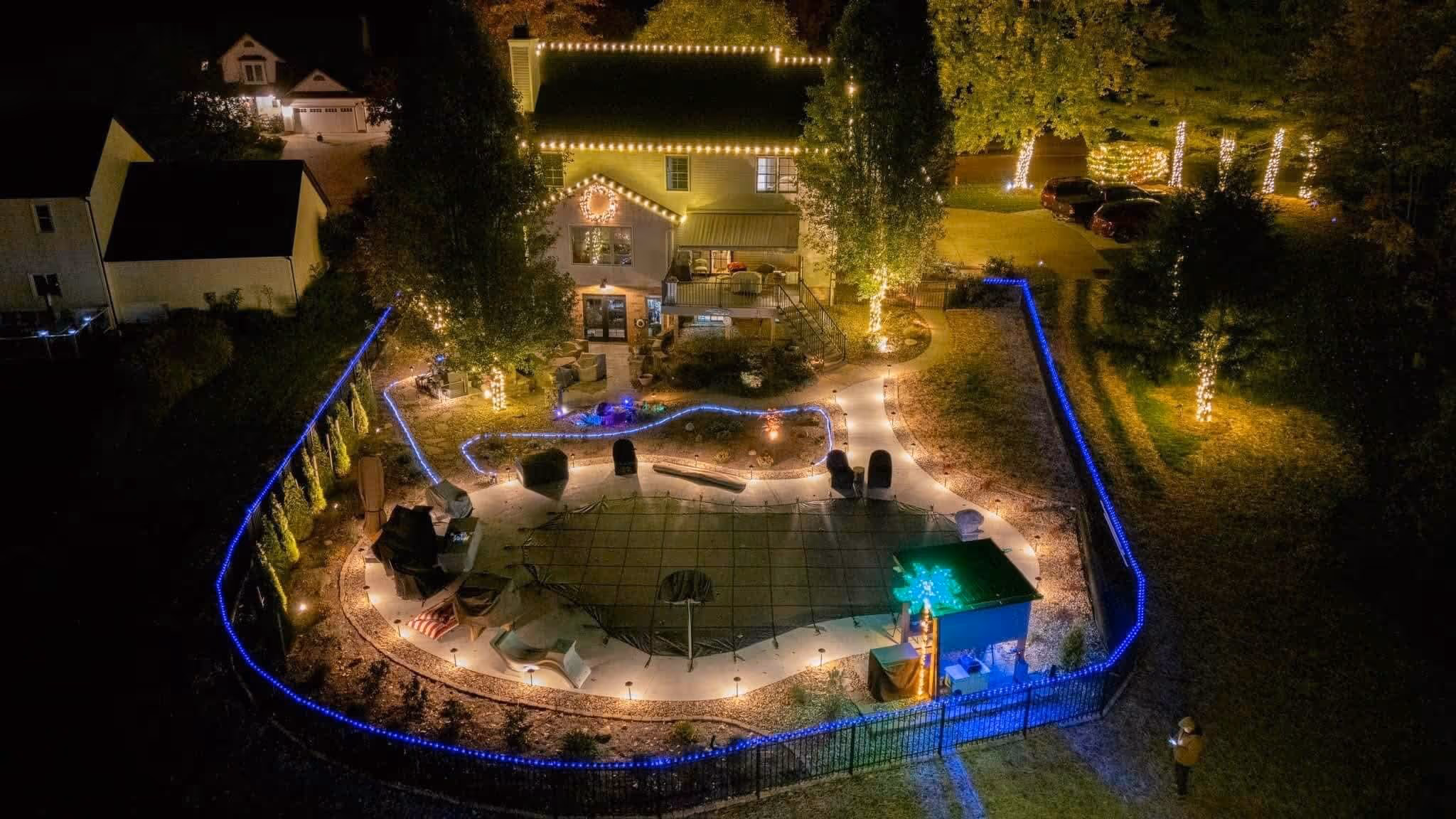 Aerial night view of a house and backyard decorated with glowing string lights on the house, trees, and blue lights outlining the backyard pool and fence.