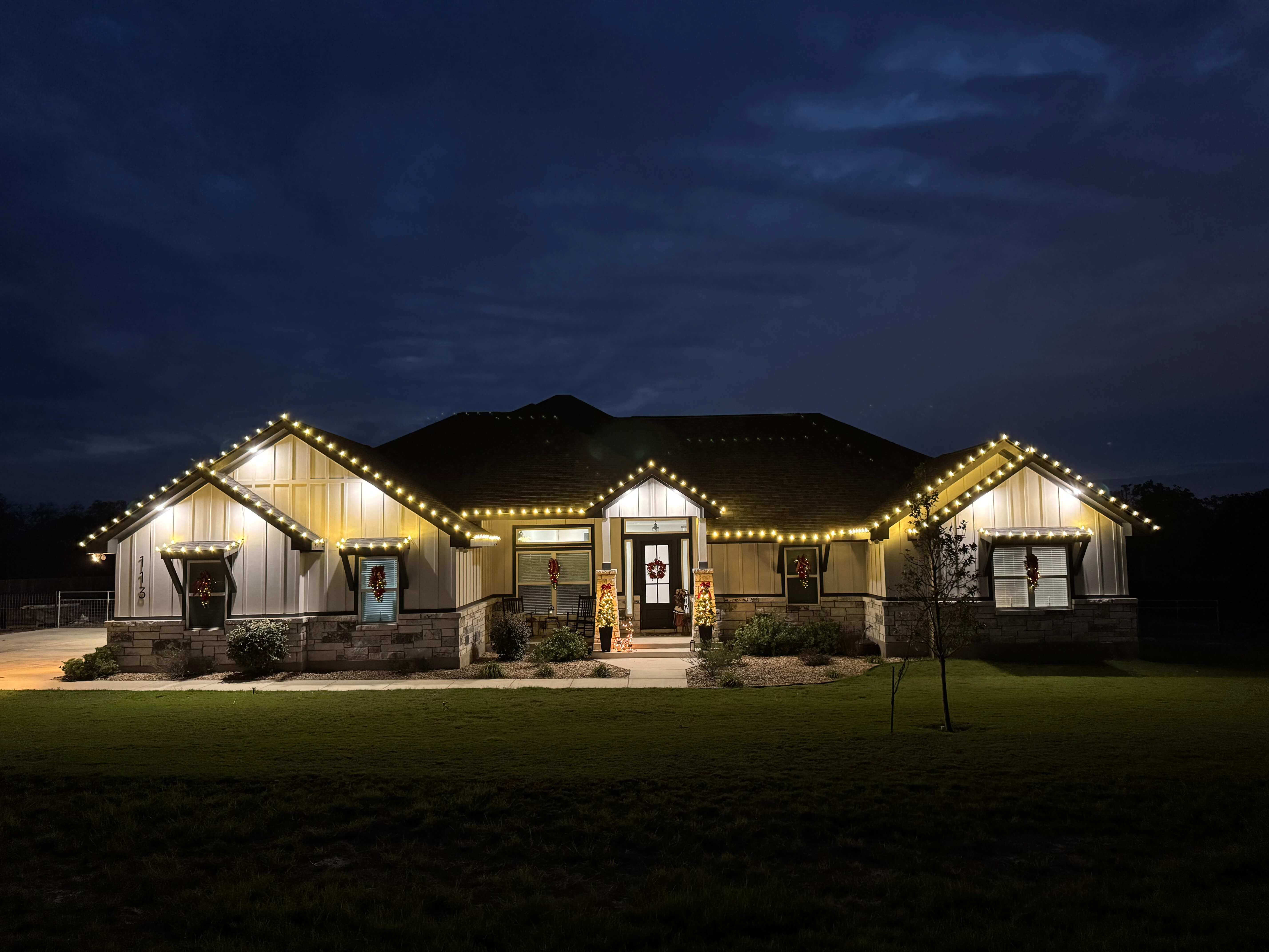 Single-story house illuminated with white string lights along roof edges and decorated with Christmas wreaths and lighted trees near the entrance at night.