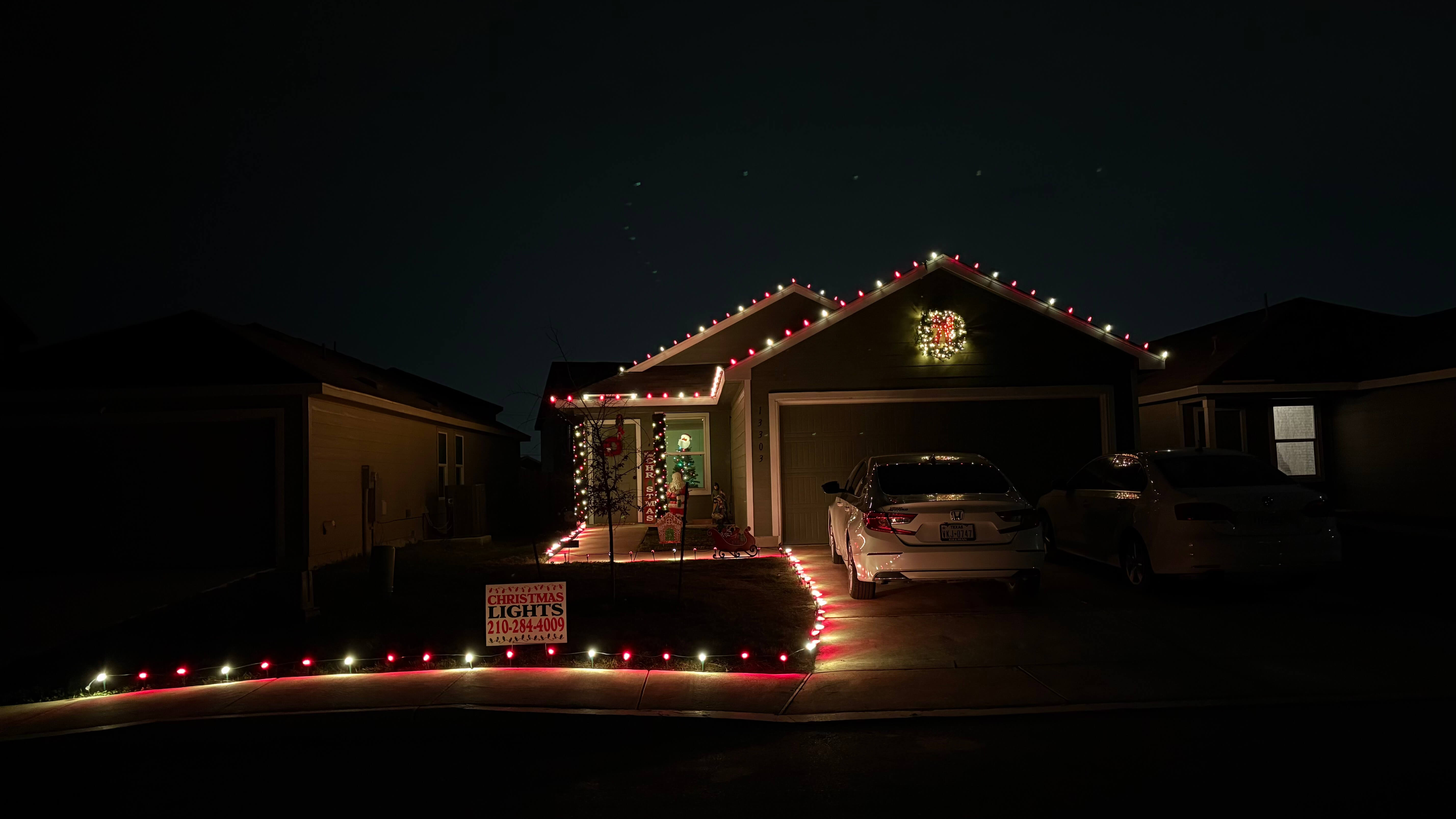 Single-story house decorated with red and white Christmas lights outlining the roof, walkway, and yard, with a lighted wreath above the garage and a sign advertising Christmas light installation services.