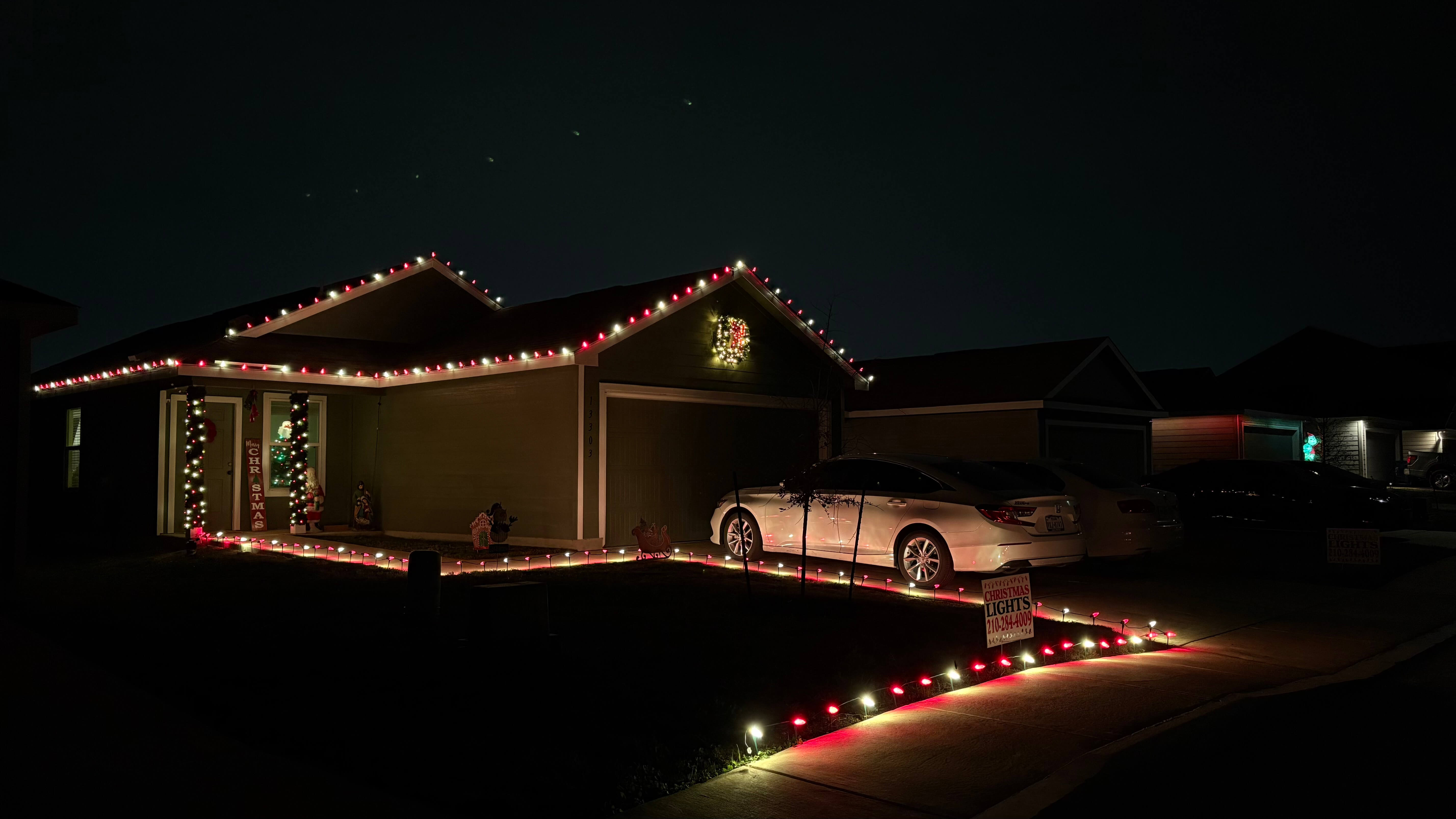House decorated with red and white Christmas lights outlining the roof, walkway, and columns, with a lighted wreath above the garage door and a white car parked in the driveway at night.