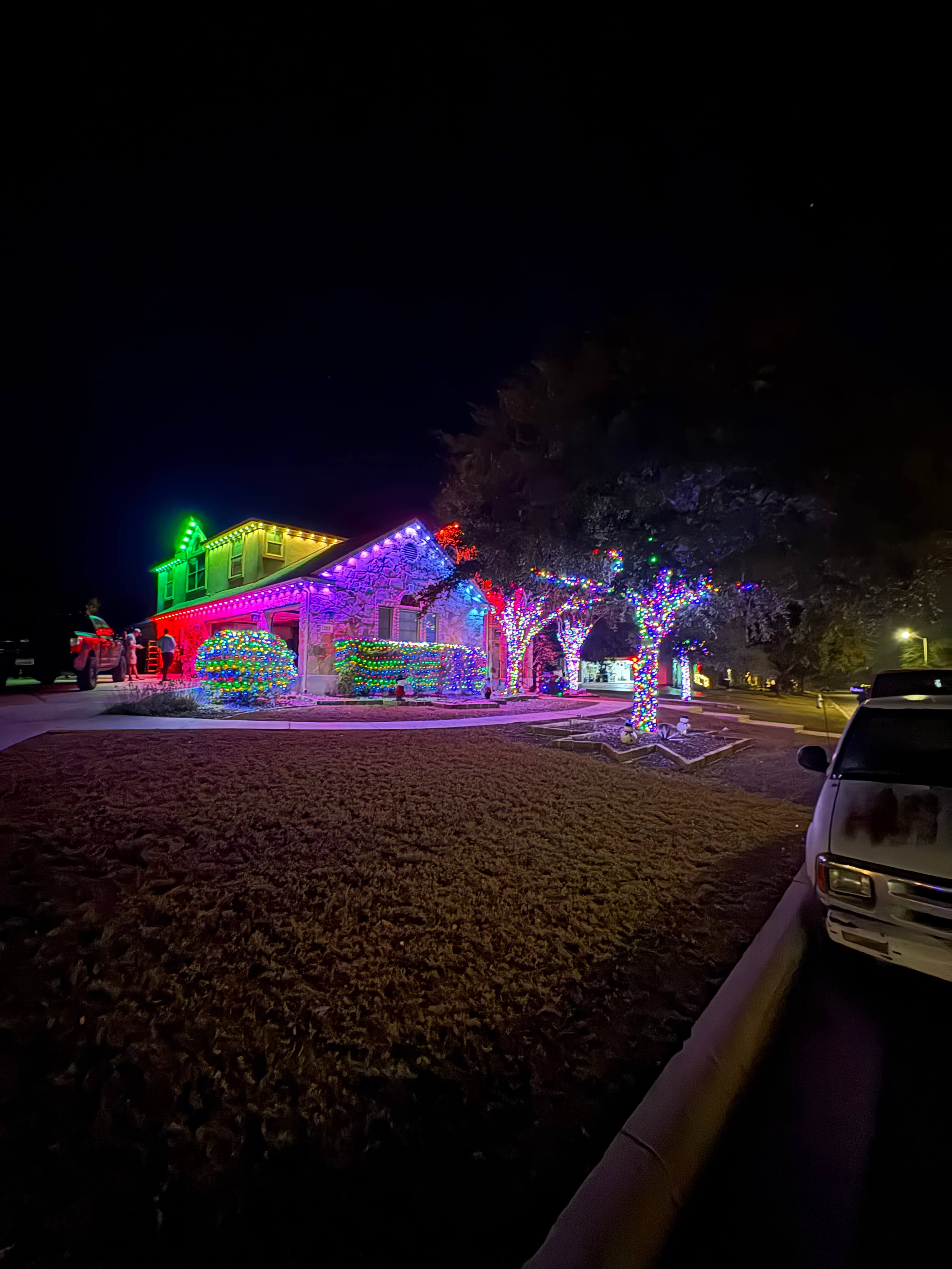 Two-story house and trees decorated with colorful Christmas lights glowing brightly at night.