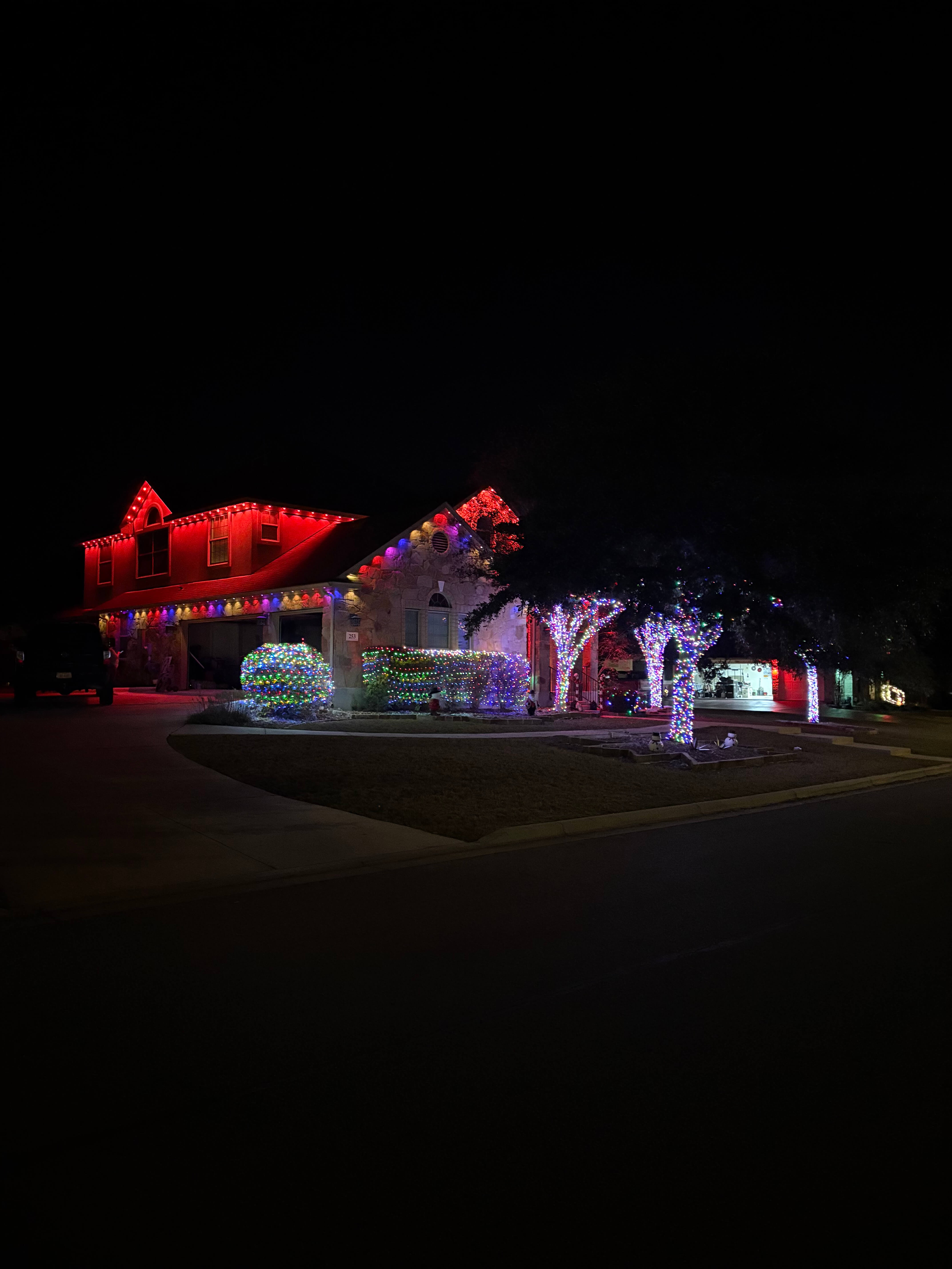 Two-story house decorated with multicolored and red Christmas lights on roof, bushes, and trees at night.