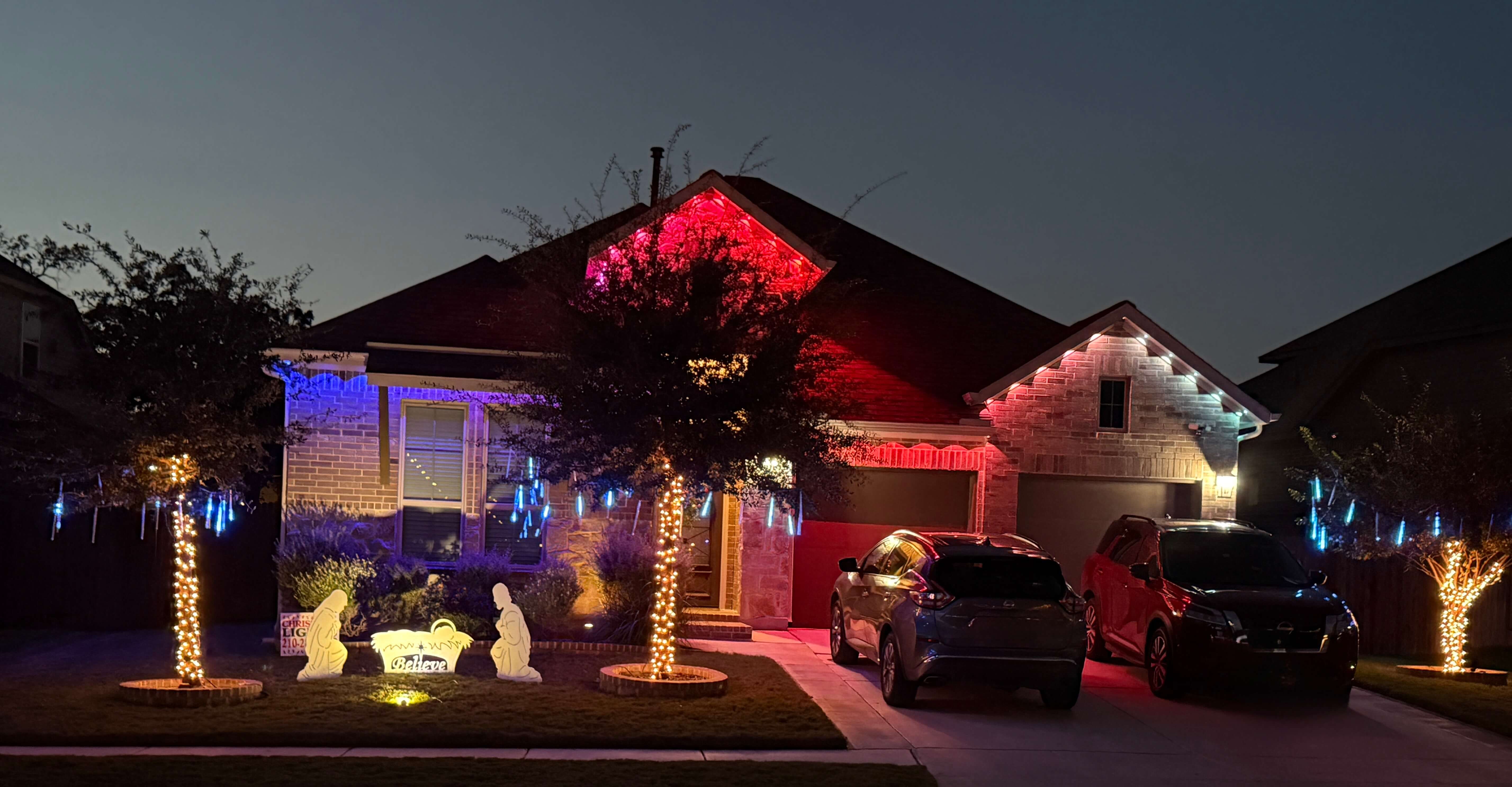 Single-story brick house at night decorated with glowing nativity scene, blue and red string lights on trees and roofline, and two cars parked in driveway.