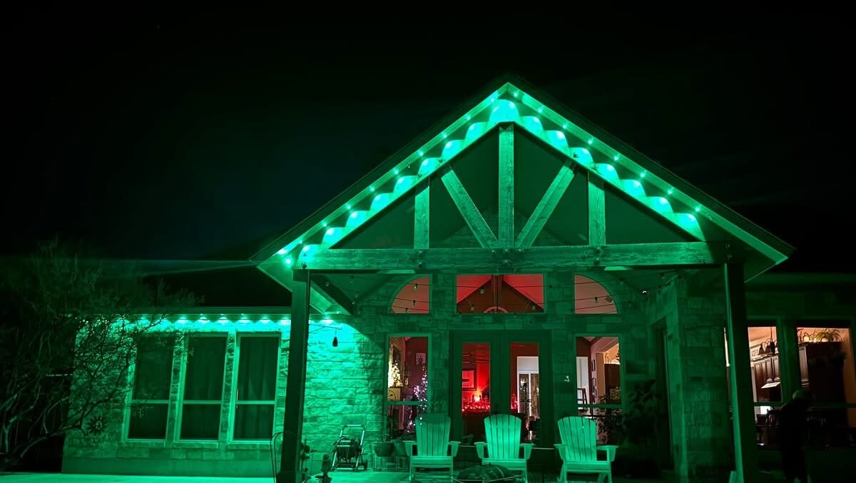 Front porch of a house illuminated with green string lights along the roofline at night, with three white chairs on the porch and warm indoor lighting visible through glass doors and windows.