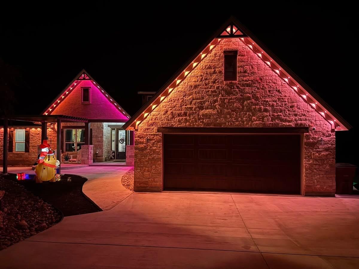 House with stone exterior and garage illuminated by pink and orange string lights at night, with inflatable holiday decorations on the left.