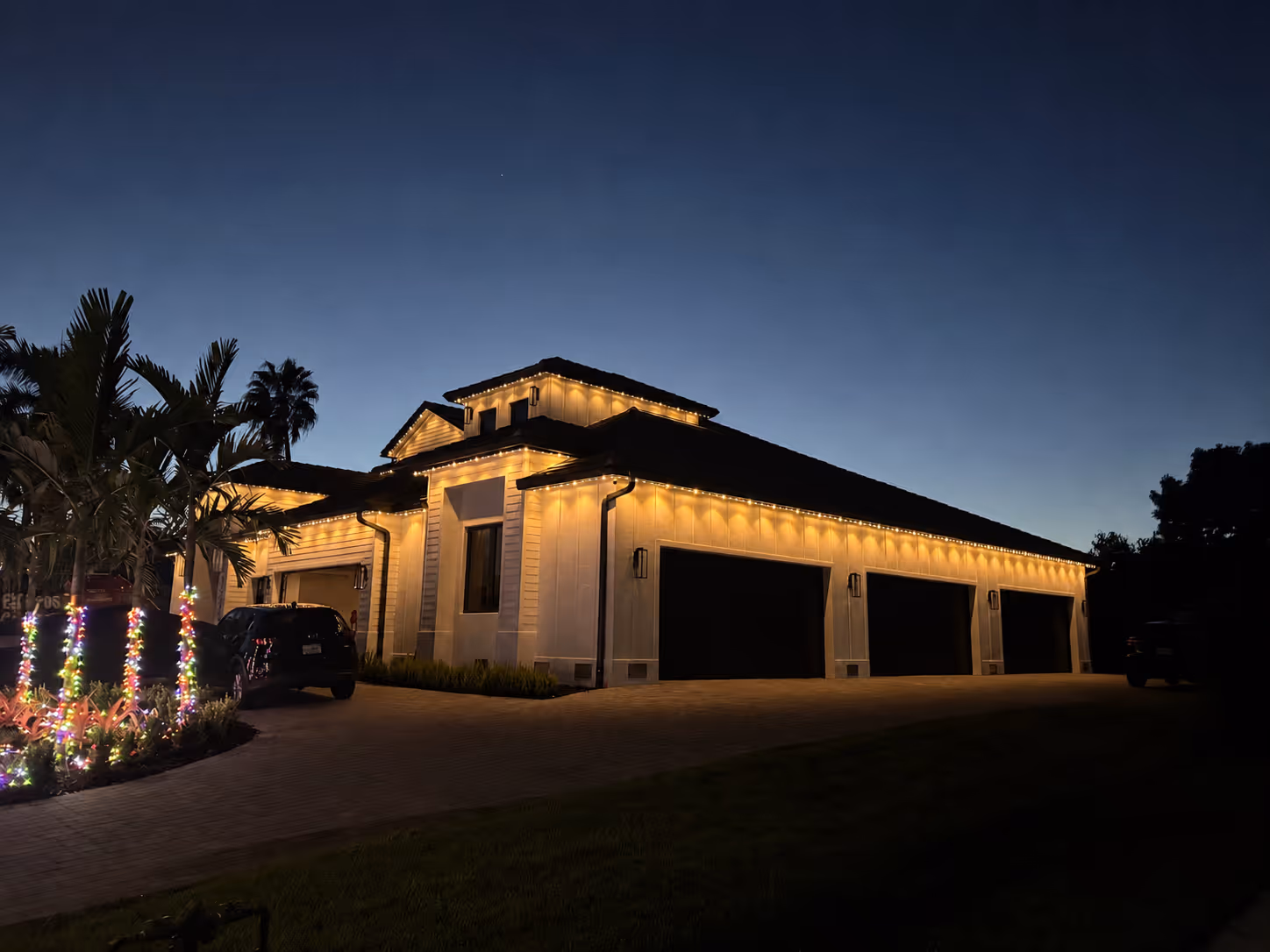 Large house at dusk with warm string lights along eaves and colorful lights wrapped around palm trees.