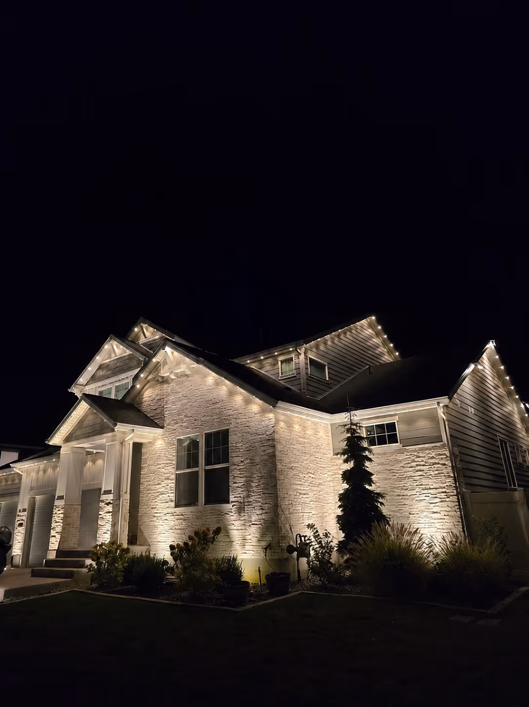 House illuminated by warm white string lights outlining roof and wall at night with garden in front.