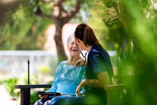 A caregiver using a stethoscope on a smiling elderly woman sitting outside on a bench surrounded by greenery.