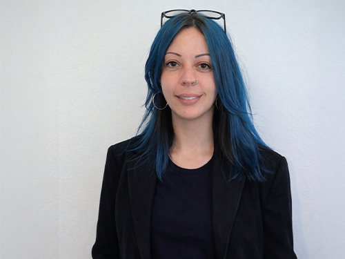 Woman with blue hair wearing glasses on her head, hoop earrings, and a black blazer over a black shirt.