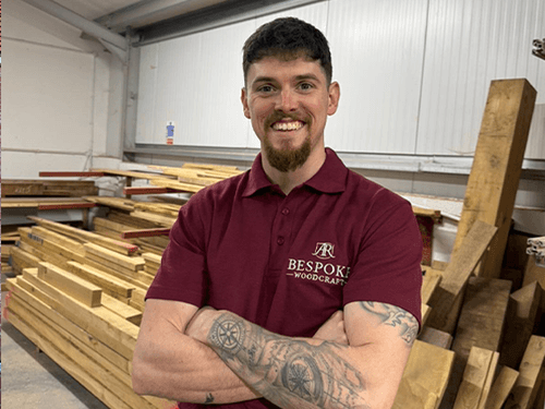 Smiling man with tattoos wearing a burgundy Bespoke Woodcraft polo in a woodshop.