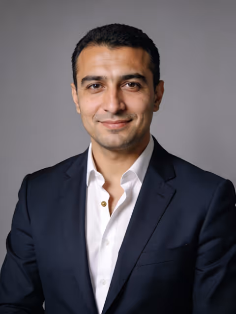 Smiling man with short dark hair wearing a navy suit jacket and white shirt against a gray background.