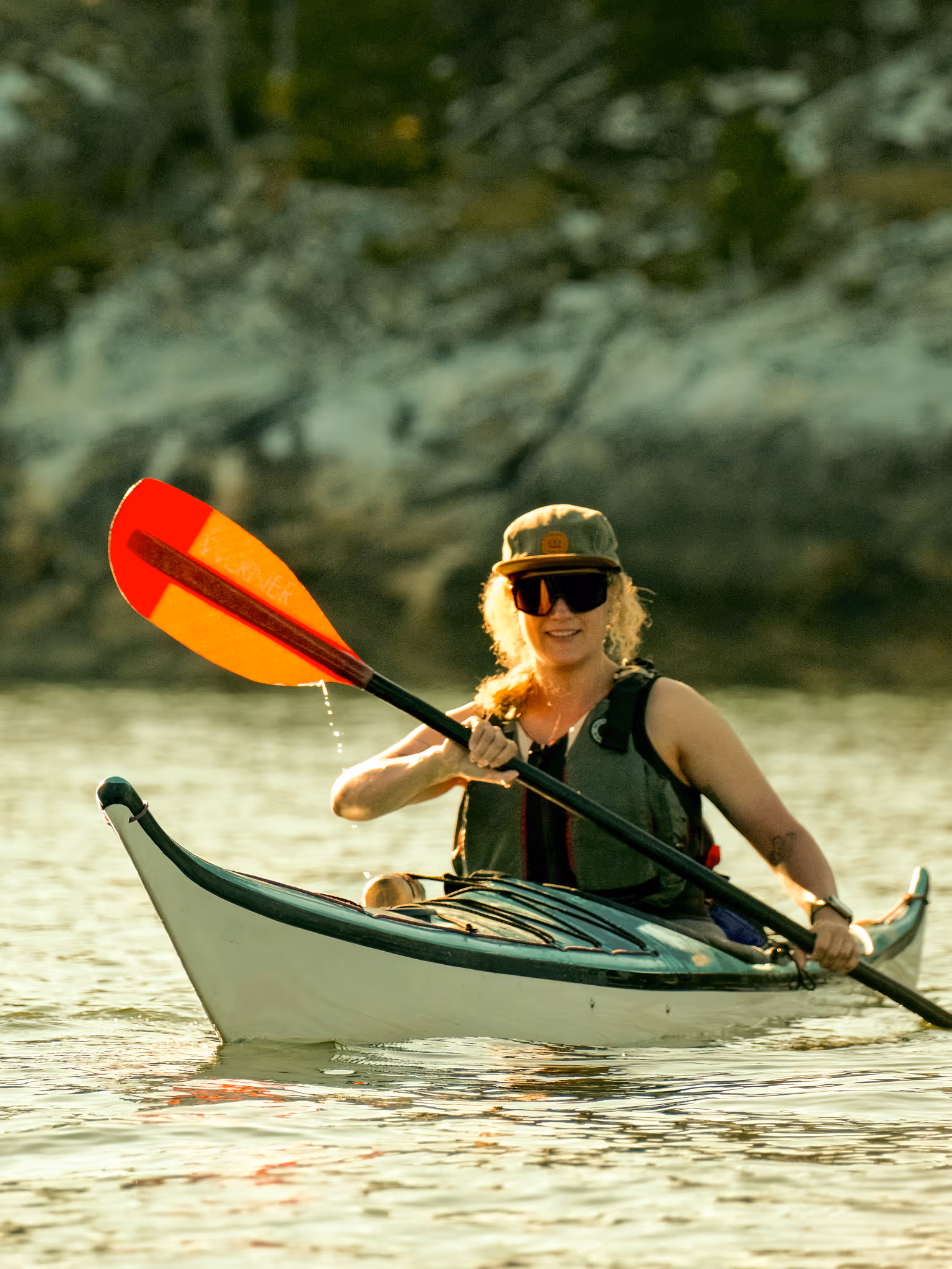 Person wearing sunglasses and a cap paddling a white kayak on calm water near a blurred shoreline.