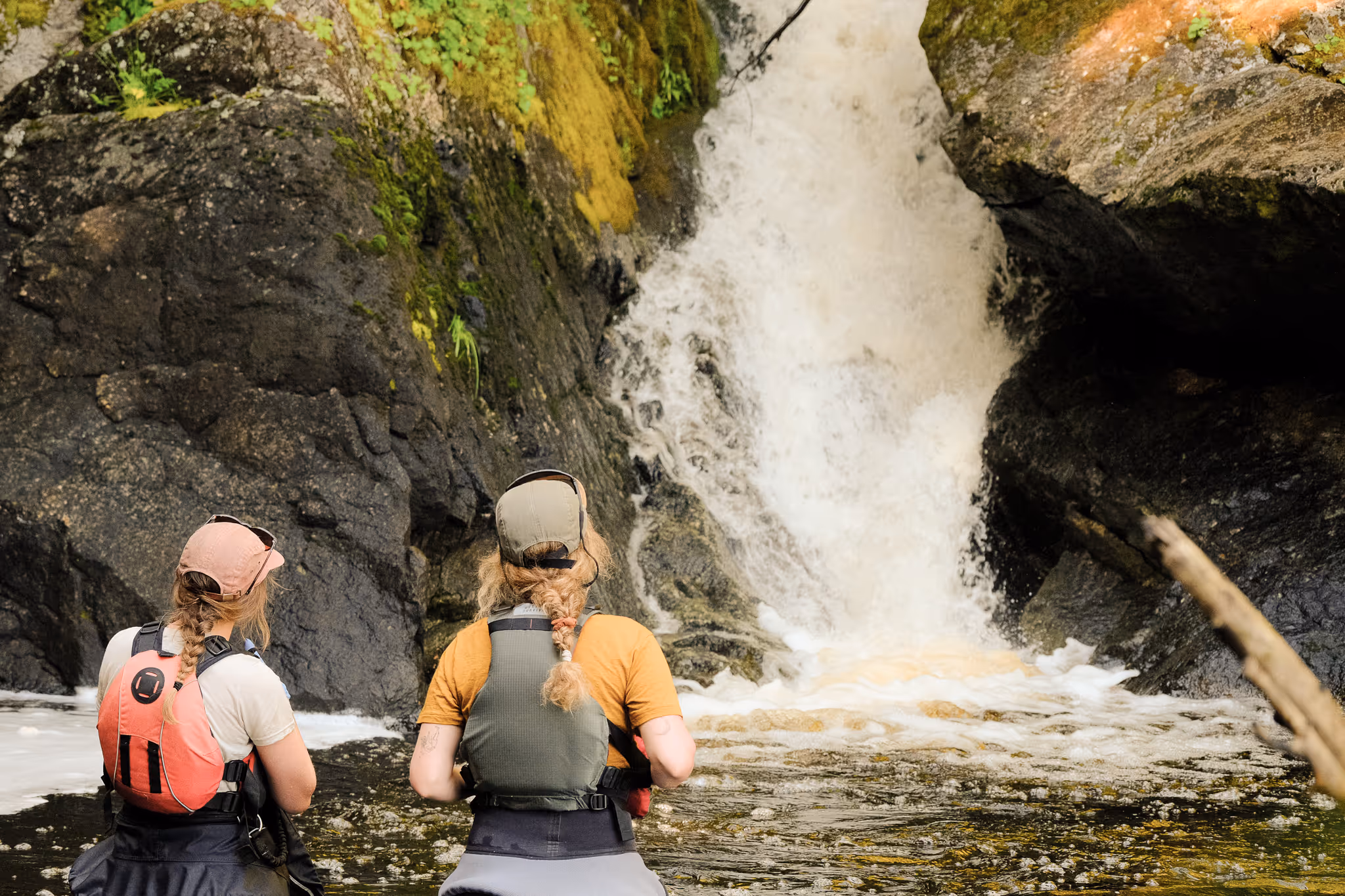 Two people in outdoor gear with braided hair facing a waterfall cascading over mossy rocks into a shallow pool.