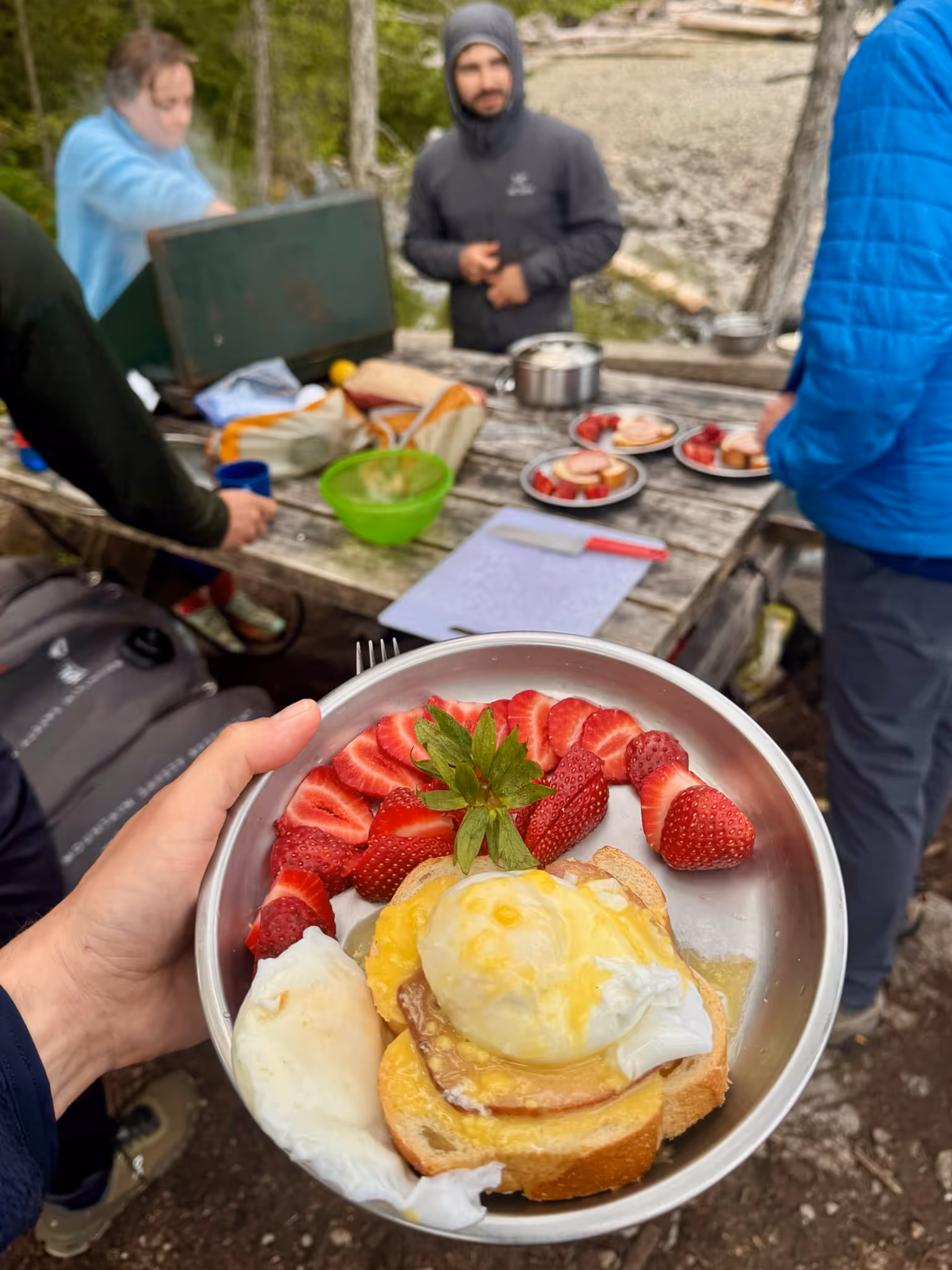 Hand holding a metal bowl with poached eggs on toast and sliced strawberries, with people preparing food on a picnic table in the background.