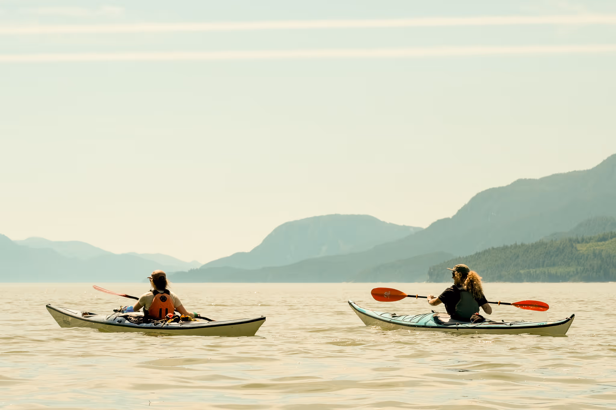 Two people kayaking on calm water with distant mountains under a clear sky.