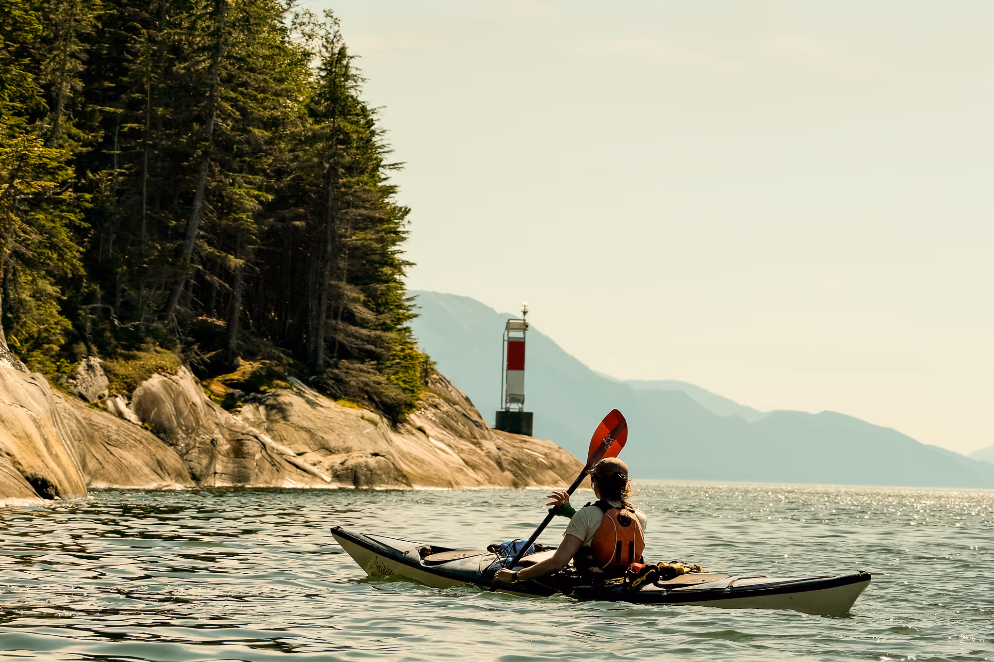 Person kayaking near rocky shore with evergreen trees and a red and white navigational marker under clear sky.