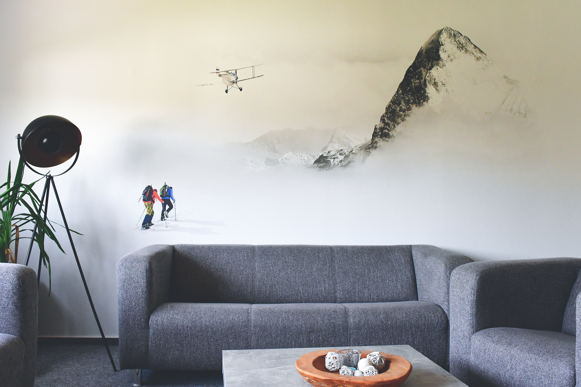 Wohnzimmer mit grauen Sofas, einem schwarzen Stehlampe-Dreibein und einer Wandmalerei von Bergsteigern, einem Flugzeug und einem schneebedeckten Berg.