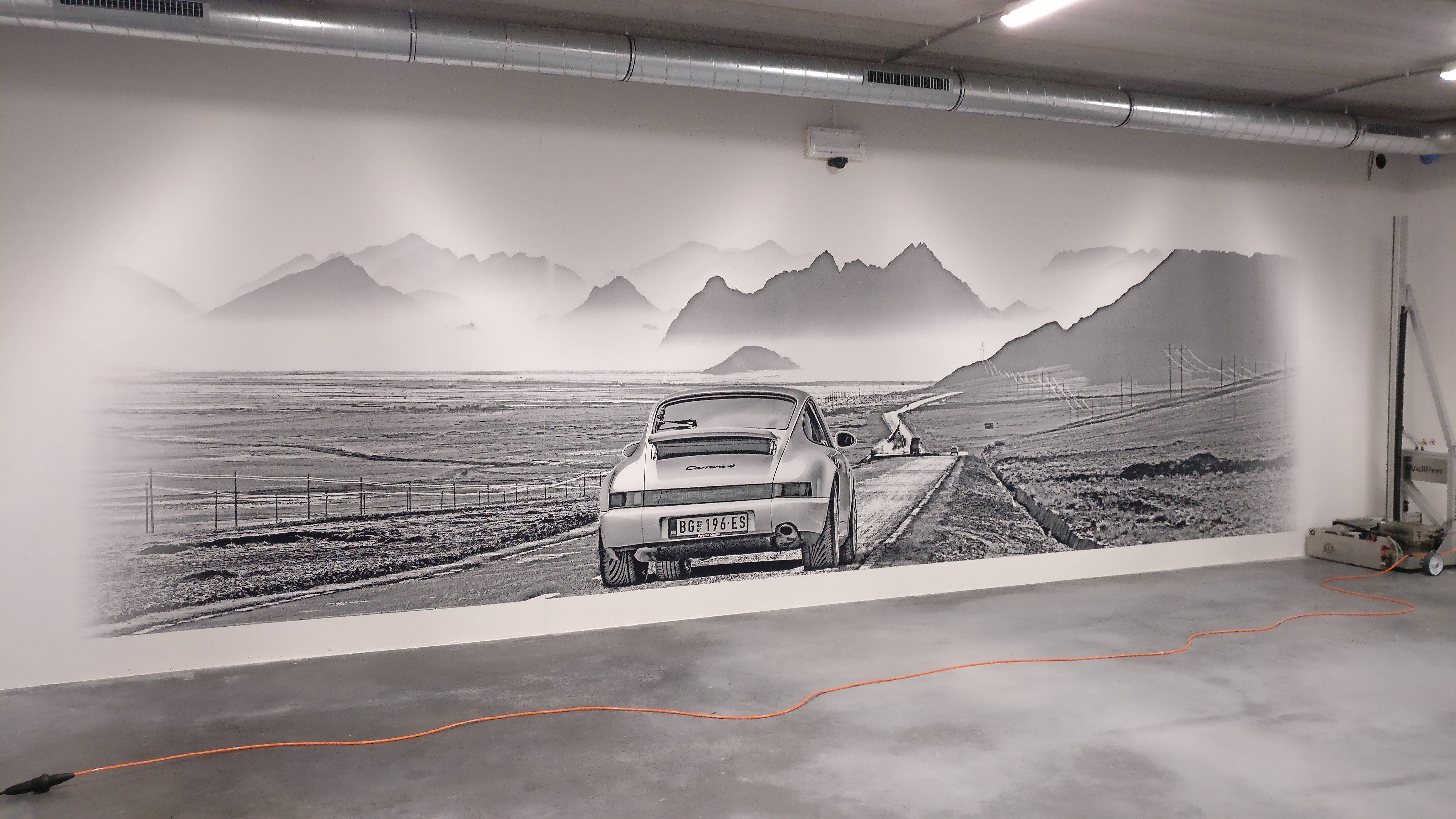 Schwarz-weißes Wandbild eines Porsche Carrera 4 auf einer Landstraße mit Bergen im Nebel im Hintergrund.