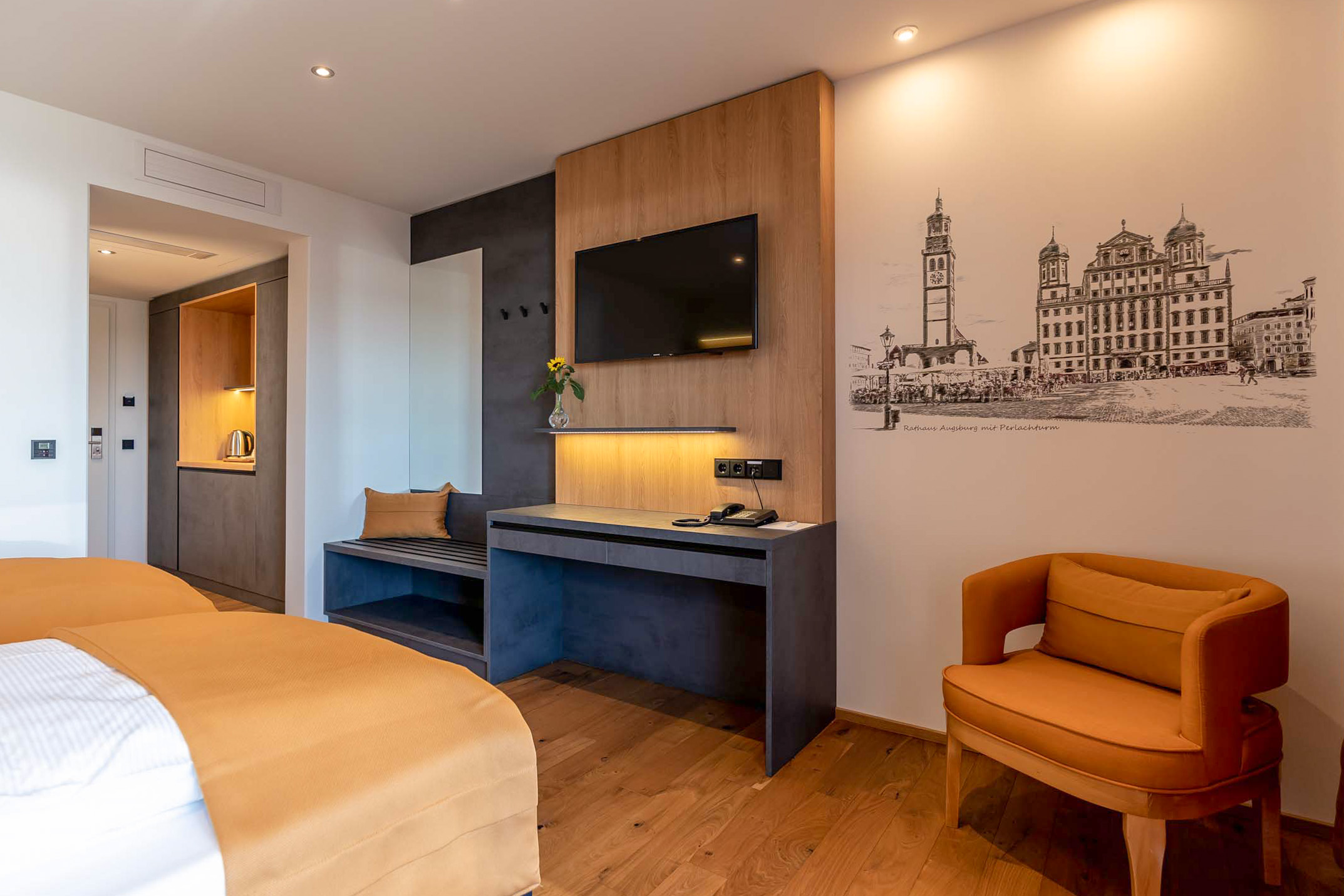 Modernes Hotelzimmer mit zwei Betten, orangefarbener Bettdecke, Wandbild von Rathaus Augsburg und gemütlichem Sessel.