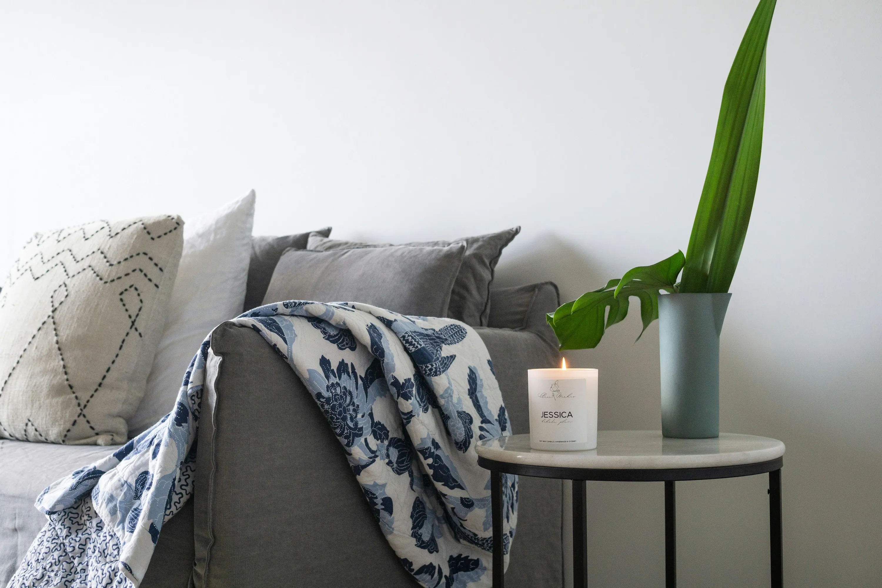Gray sofa with patterned cushions and a blue floral throw blanket next to a side table with a lit candle and a green plant in a vase.