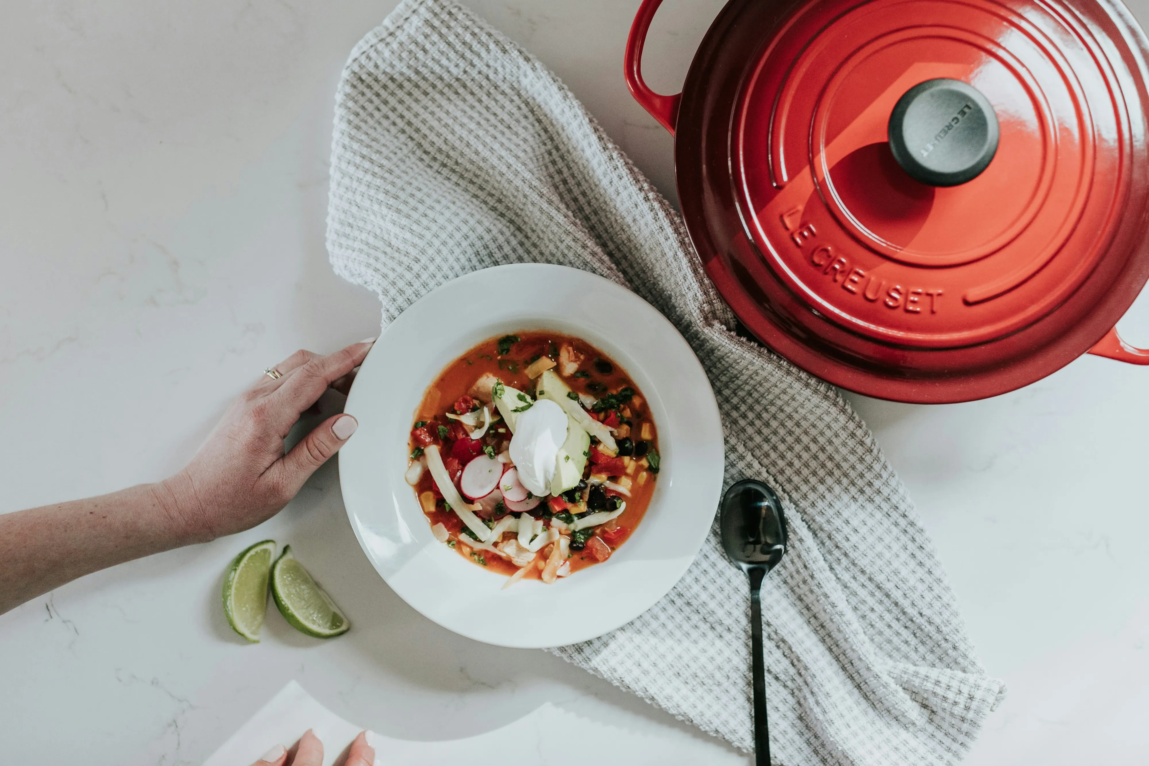 Top-down view of a bowl of vegetable soup with radishes and sour cream, a red Le Creuset pot, a black spoon, lime wedges, and a hand on a white countertop.