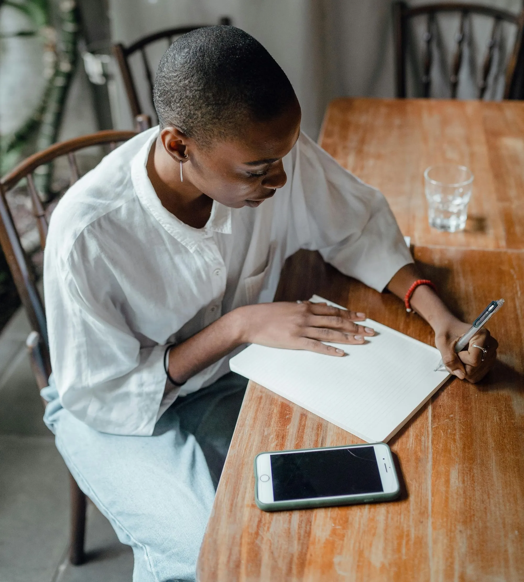Person with short hair in a white shirt writing in a notebook at a wooden table with a smartphone and a glass of water nearby.