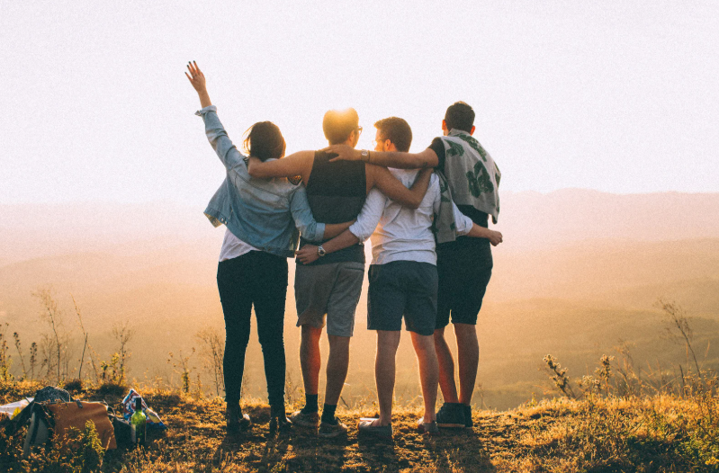 Group of friends with arms around each other looking at a sunset over a valley after a hike.
